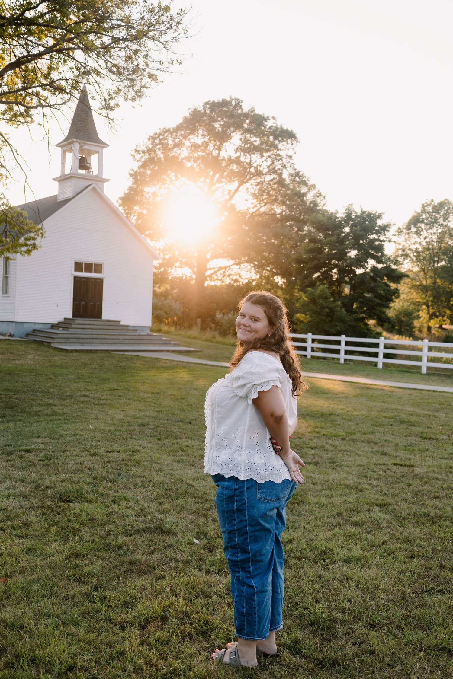 Young woman in white top and jeans smiling outdoors at golden hour near a white country church with steeple.