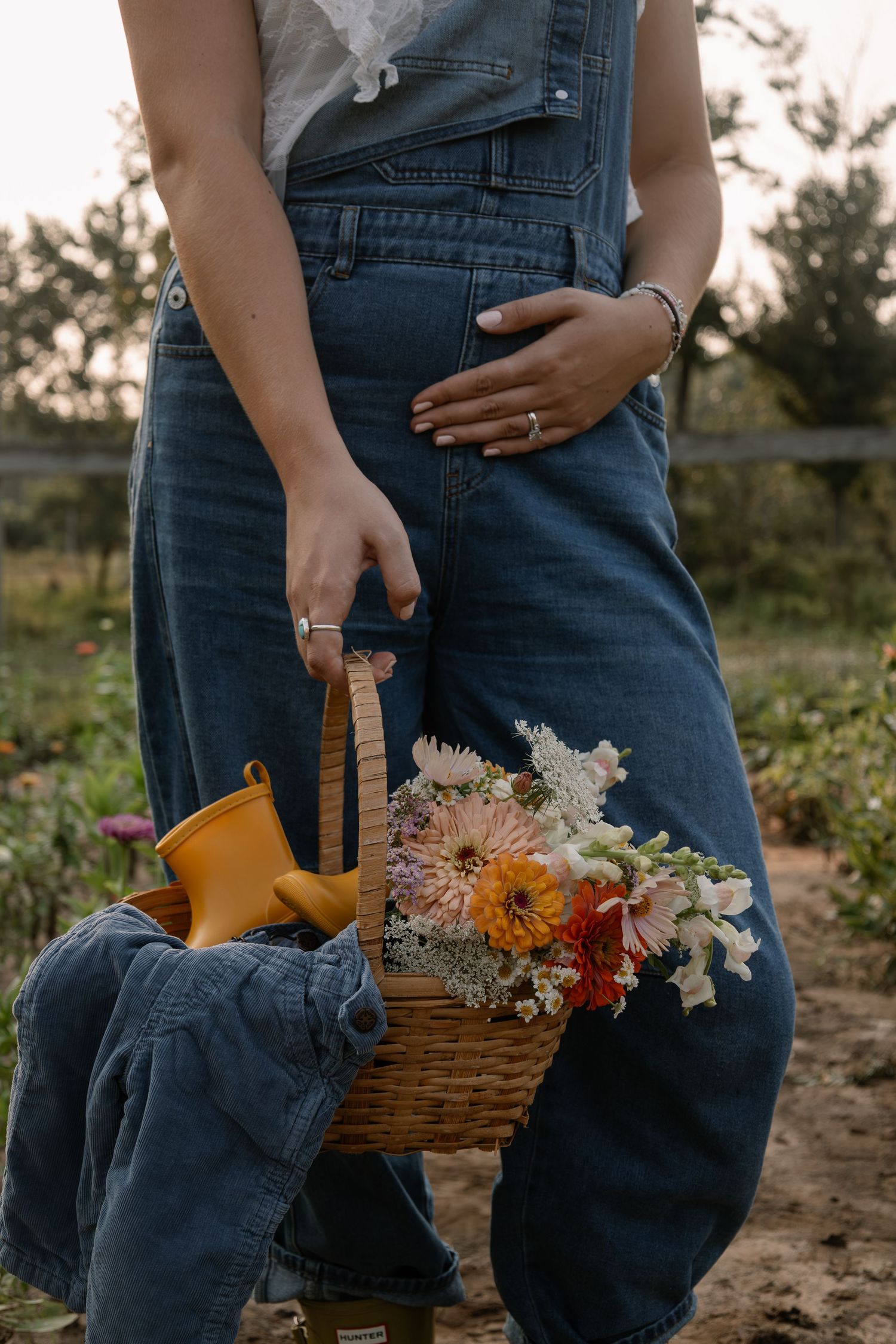 Pregnant woman in denim overalls holding a wicker basket with fresh flowers and yellow rain boots in a garden.