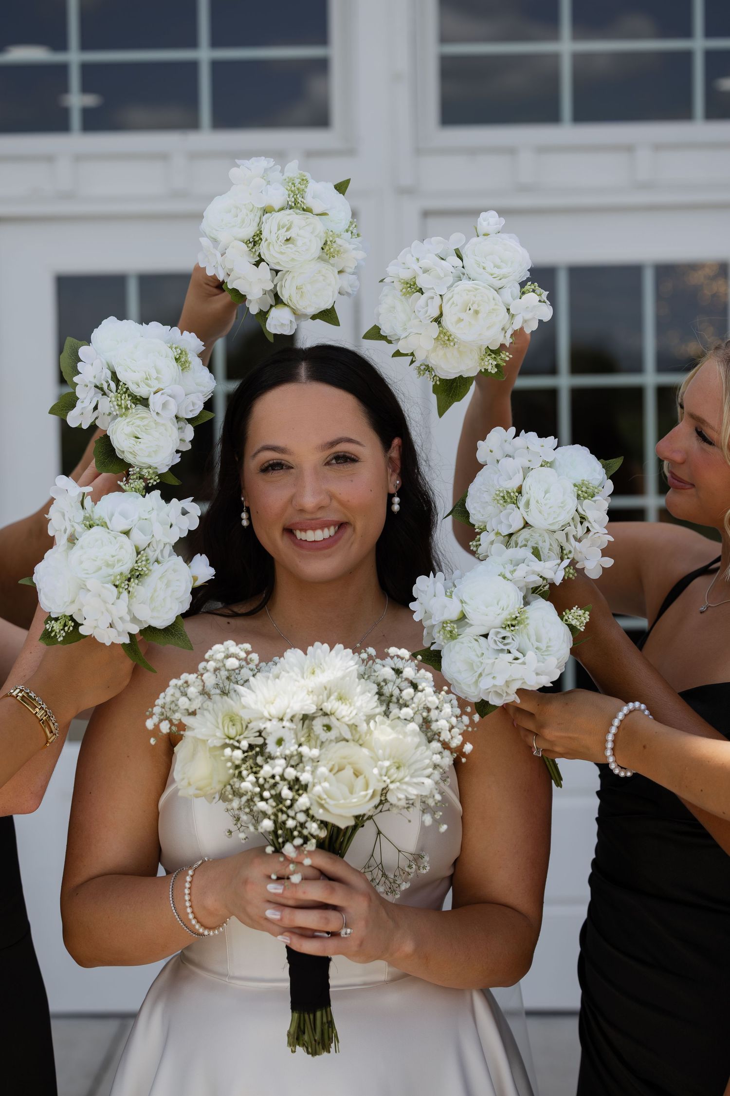 Smiling bride in white gown holds bouquet of white roses and baby's breath, surrounded by bridesmaids raising their bouquets.