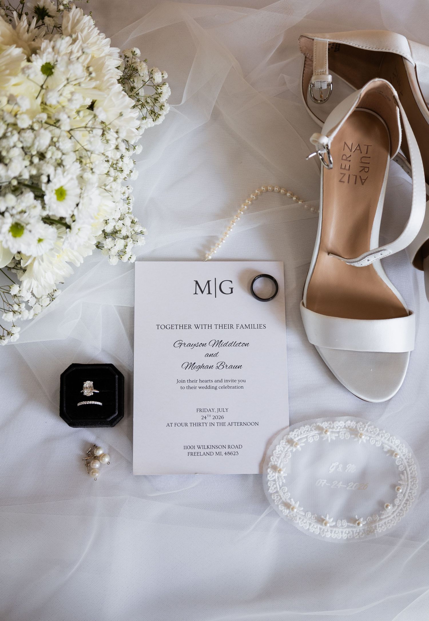 Elegant wedding flat lay with white heels, floral bouquet, pearl necklace, engagement ring, and monogram invitation on white fabric.