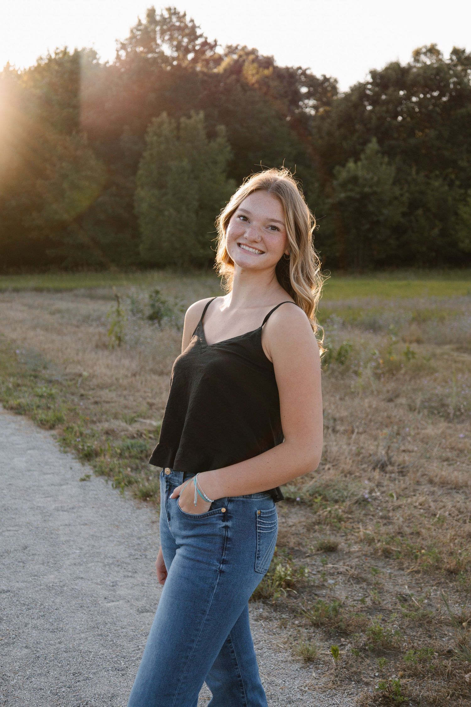 Smiling teenage girl in black tank top and jeans poses on a path during golden hour sunset outdoors.