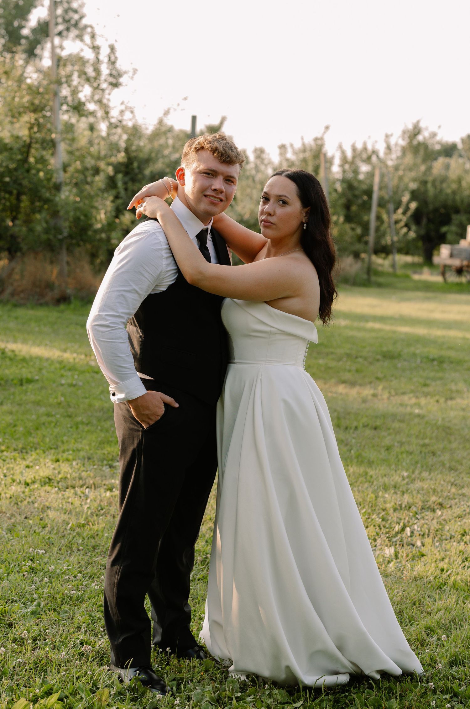 Bride in strapless white gown embracing groom in black vest outdoors at sunset in a green orchard.