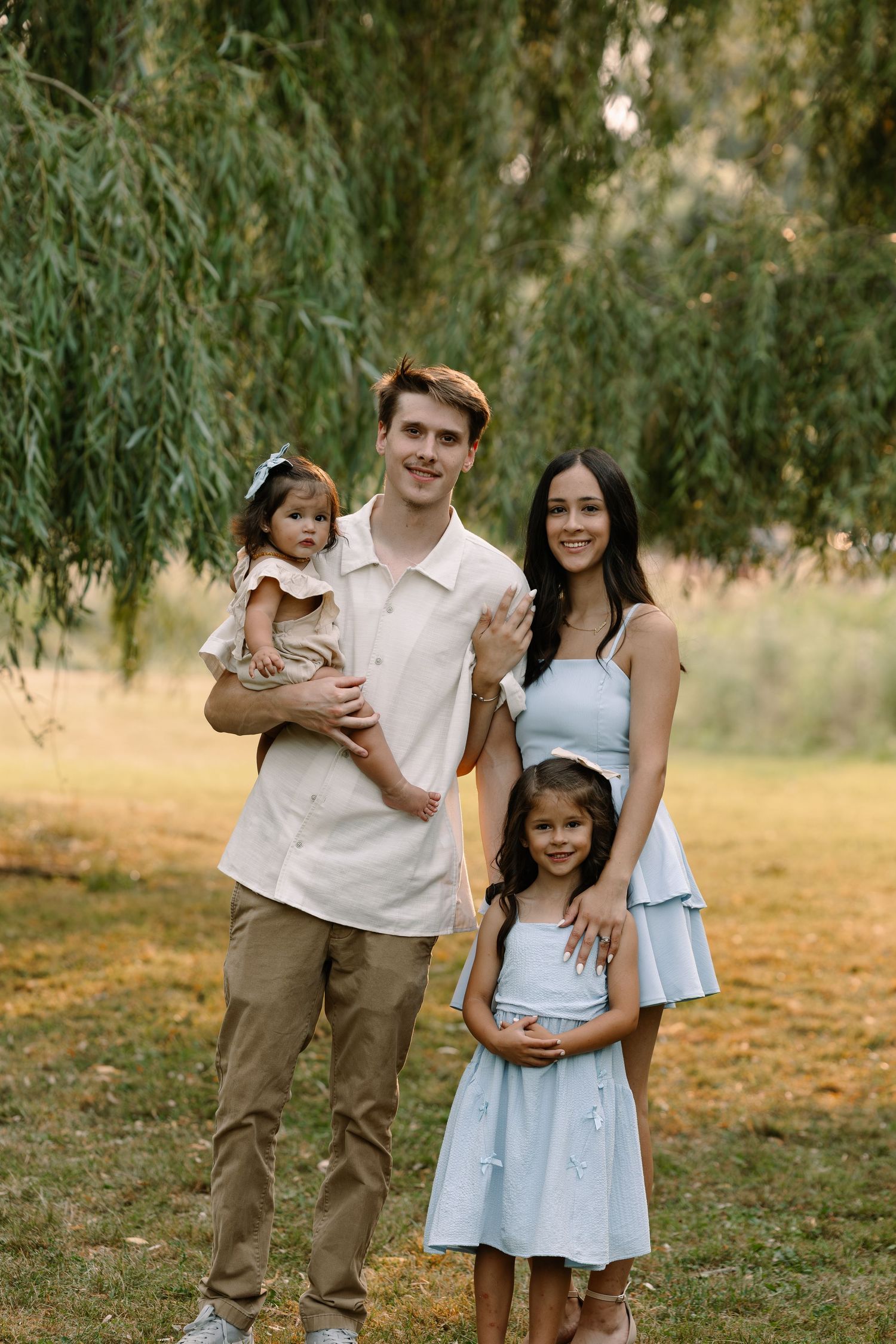 Young family of four posing outdoors under willow tree, parents with two daughters in light blue dresses on green lawn.