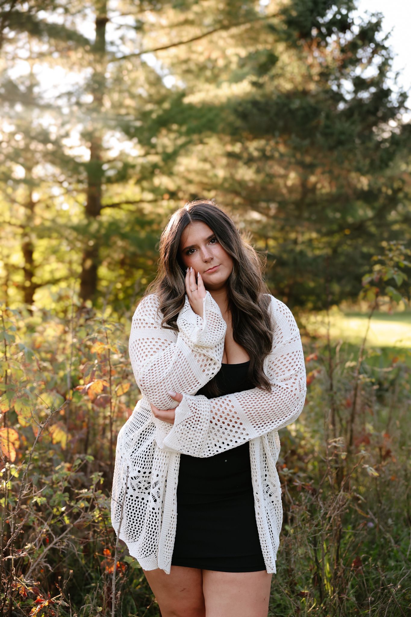 Young woman in white knit cardigan and black skirt posing in an autumn forest setting.