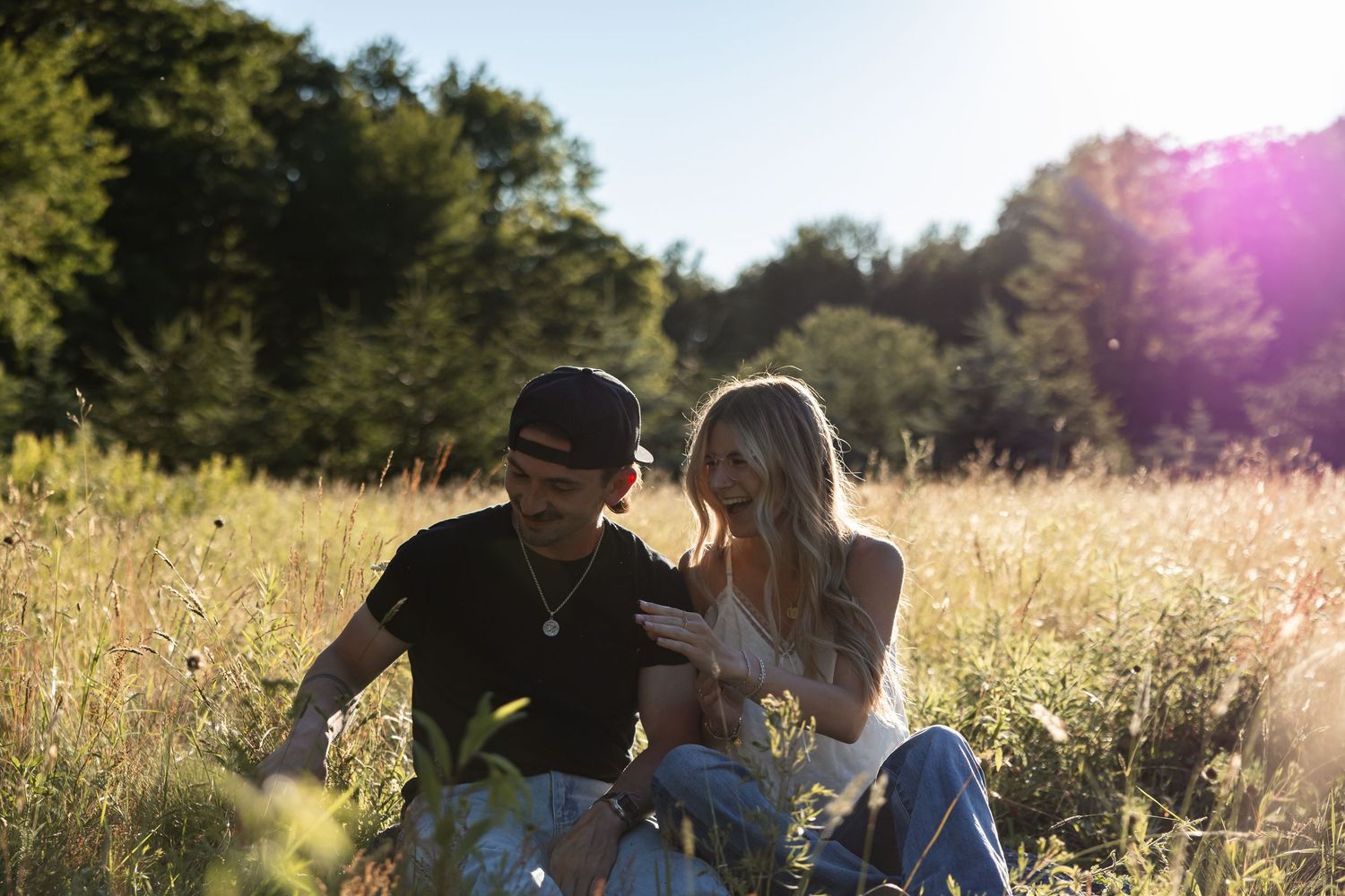 Couple sitting close together in a bright sunlit field, sharing an intimate moment among wildflowers and greenery.