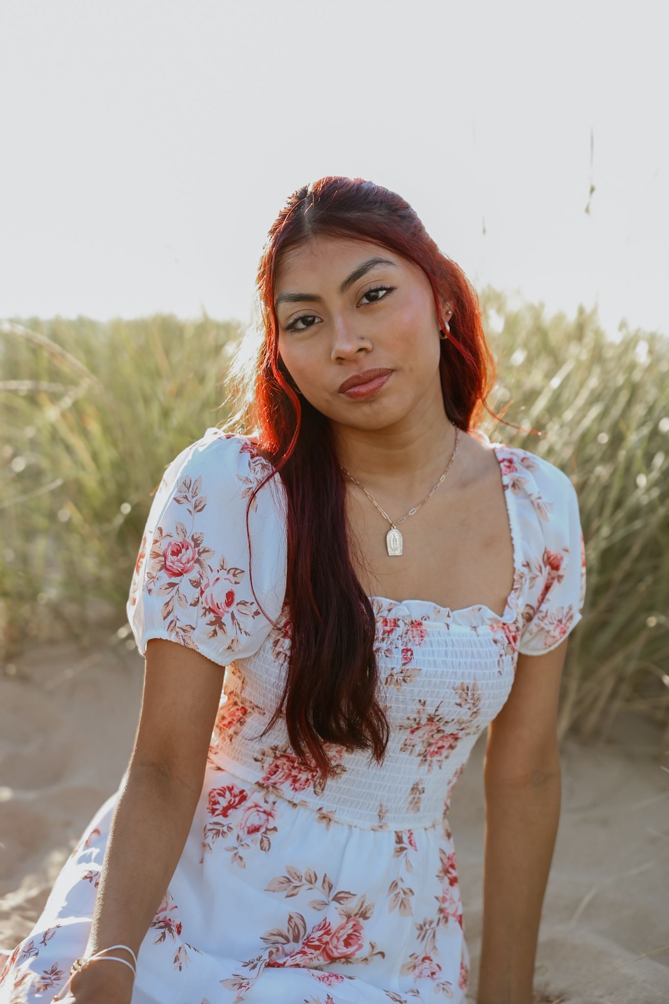 Young woman in floral dress sitting on sandy beach at sunset.