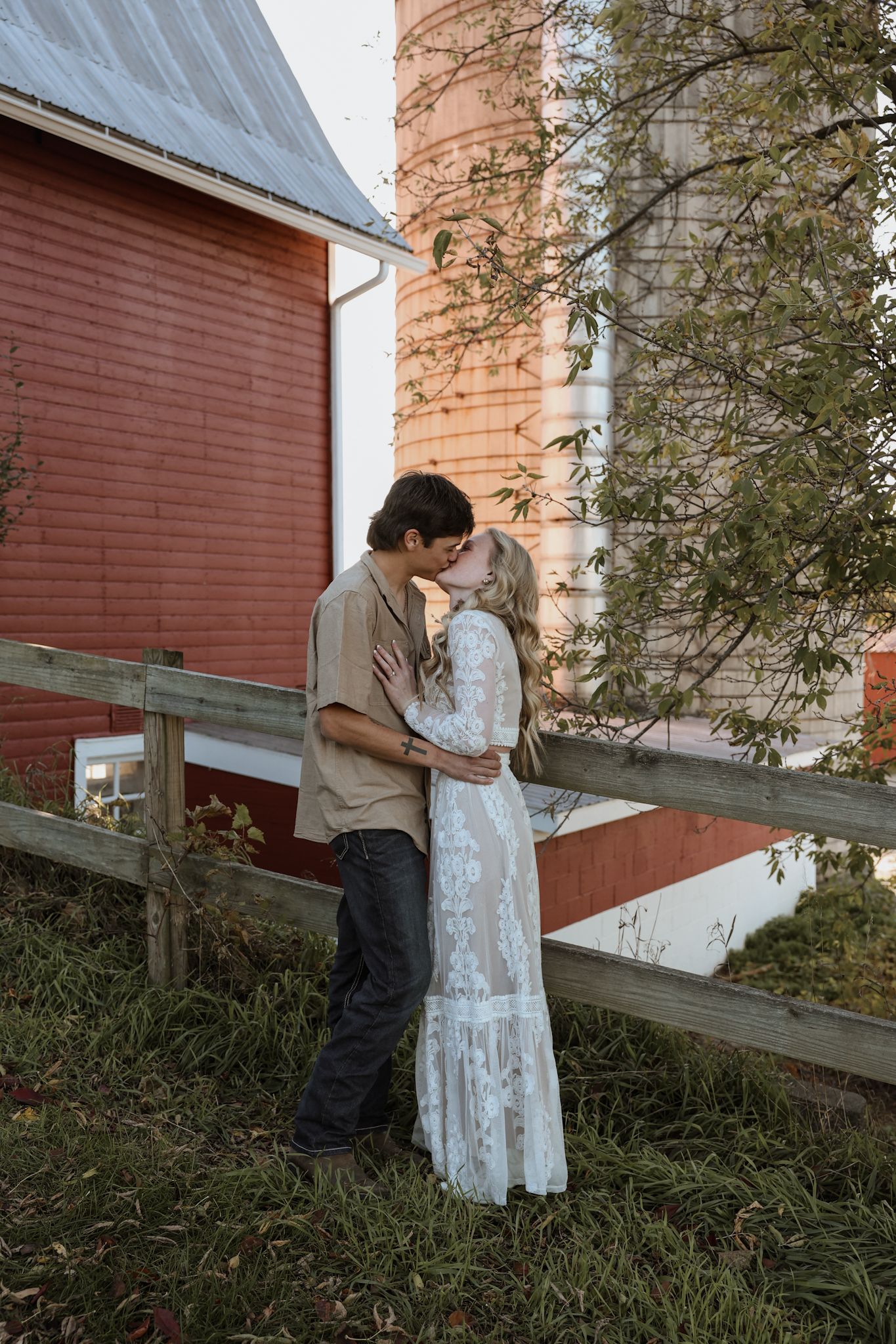 Romantic couple sharing a kiss near a wooden fence with a red barn and silo backdrop.