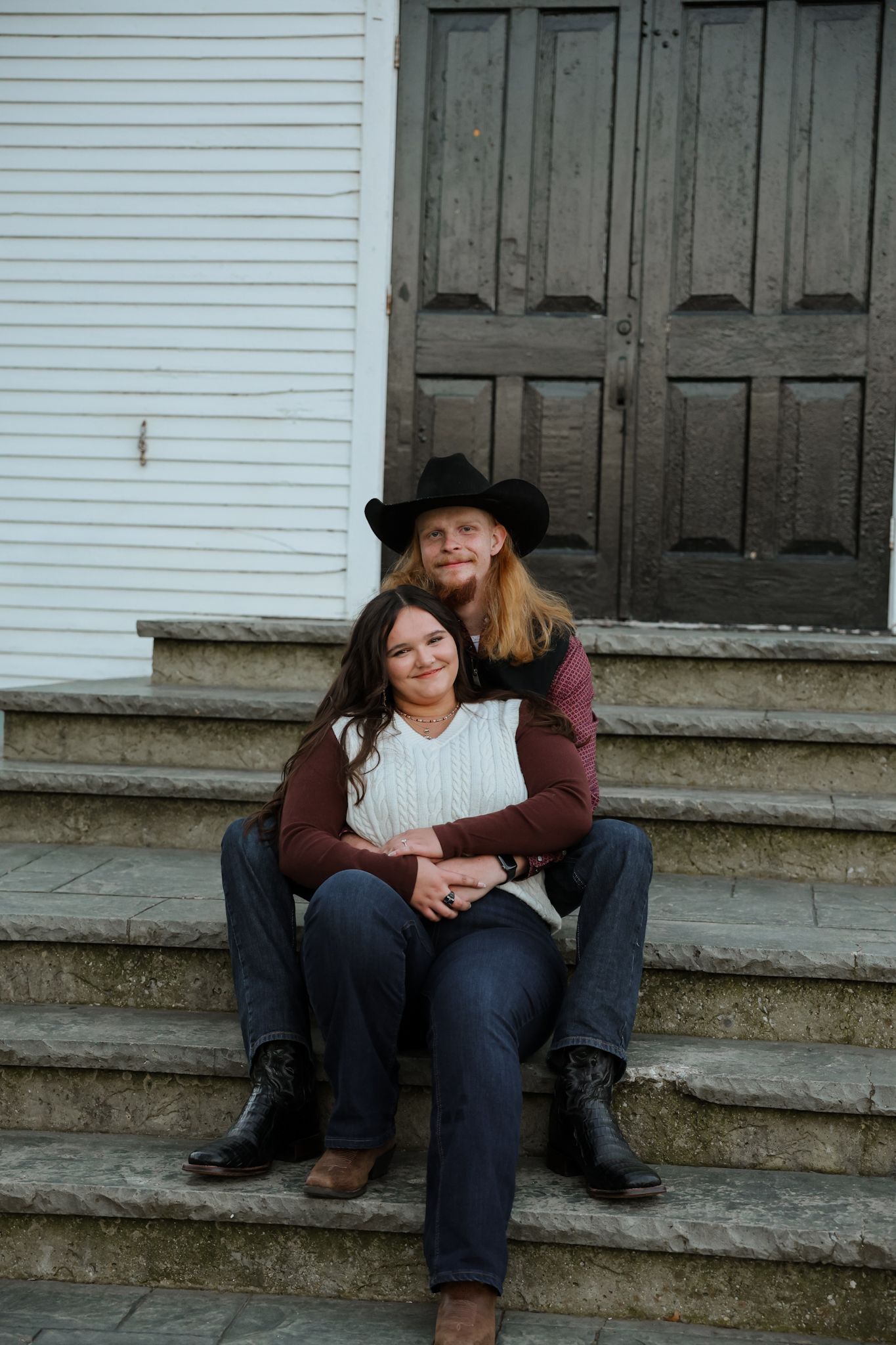 Couple sitting on stone steps embracing, man in black cowboy hat, fall setting.