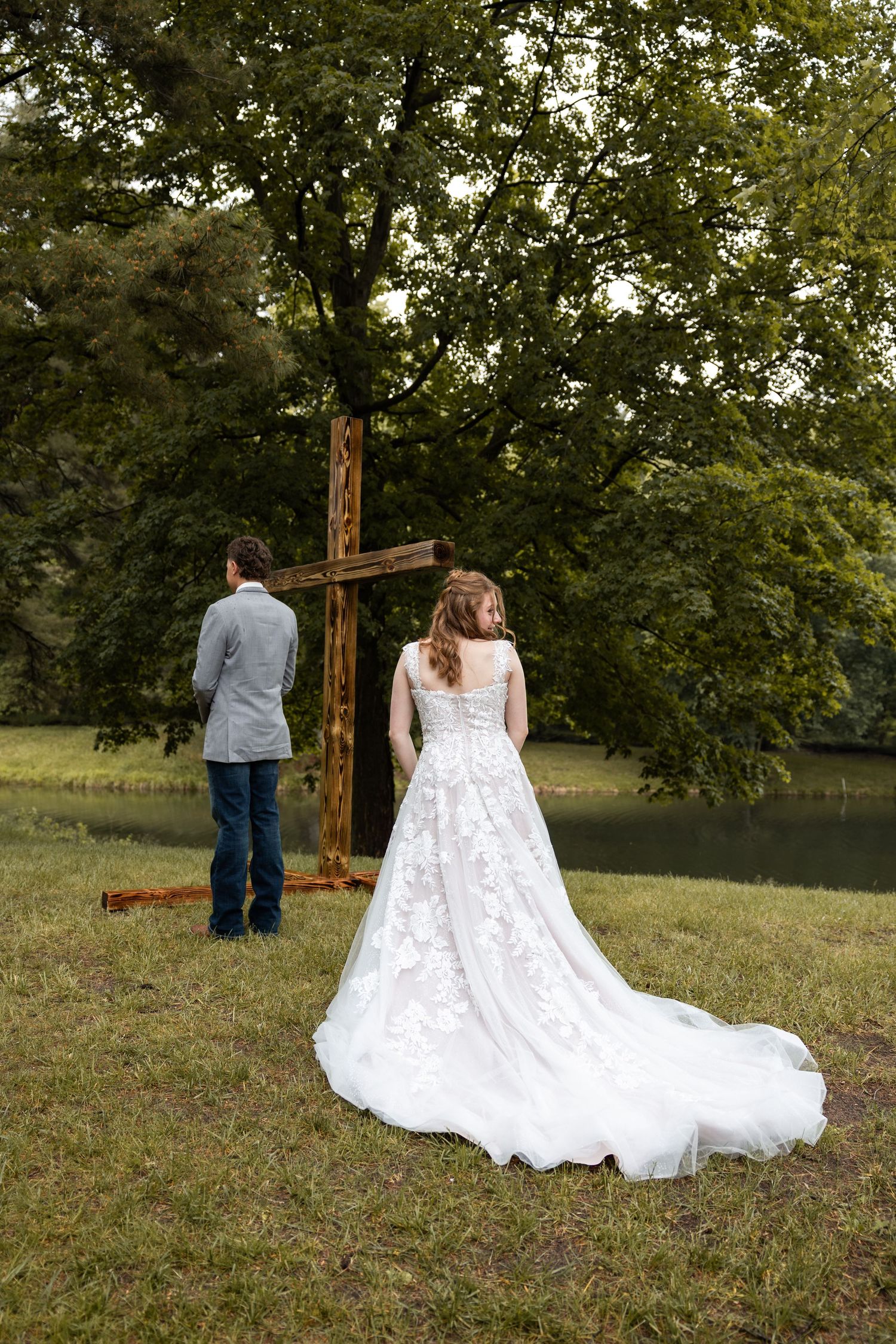 Bride and groom stand before a rustic wooden cross outdoors near a tree-lined pond.