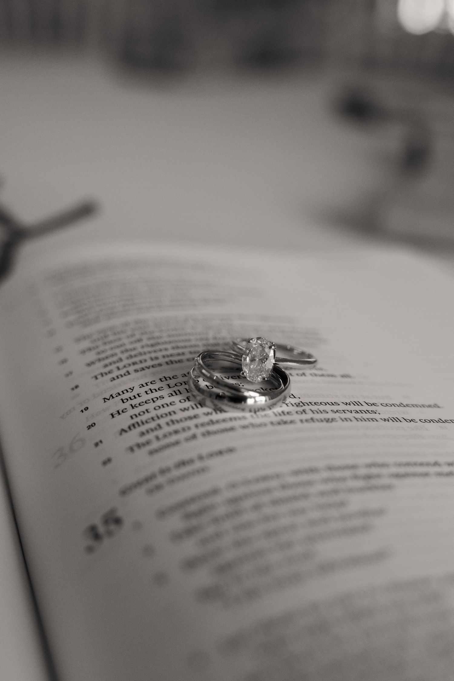 Black and white close-up of diamond engagement ring and wedding bands resting on open Bible pages.