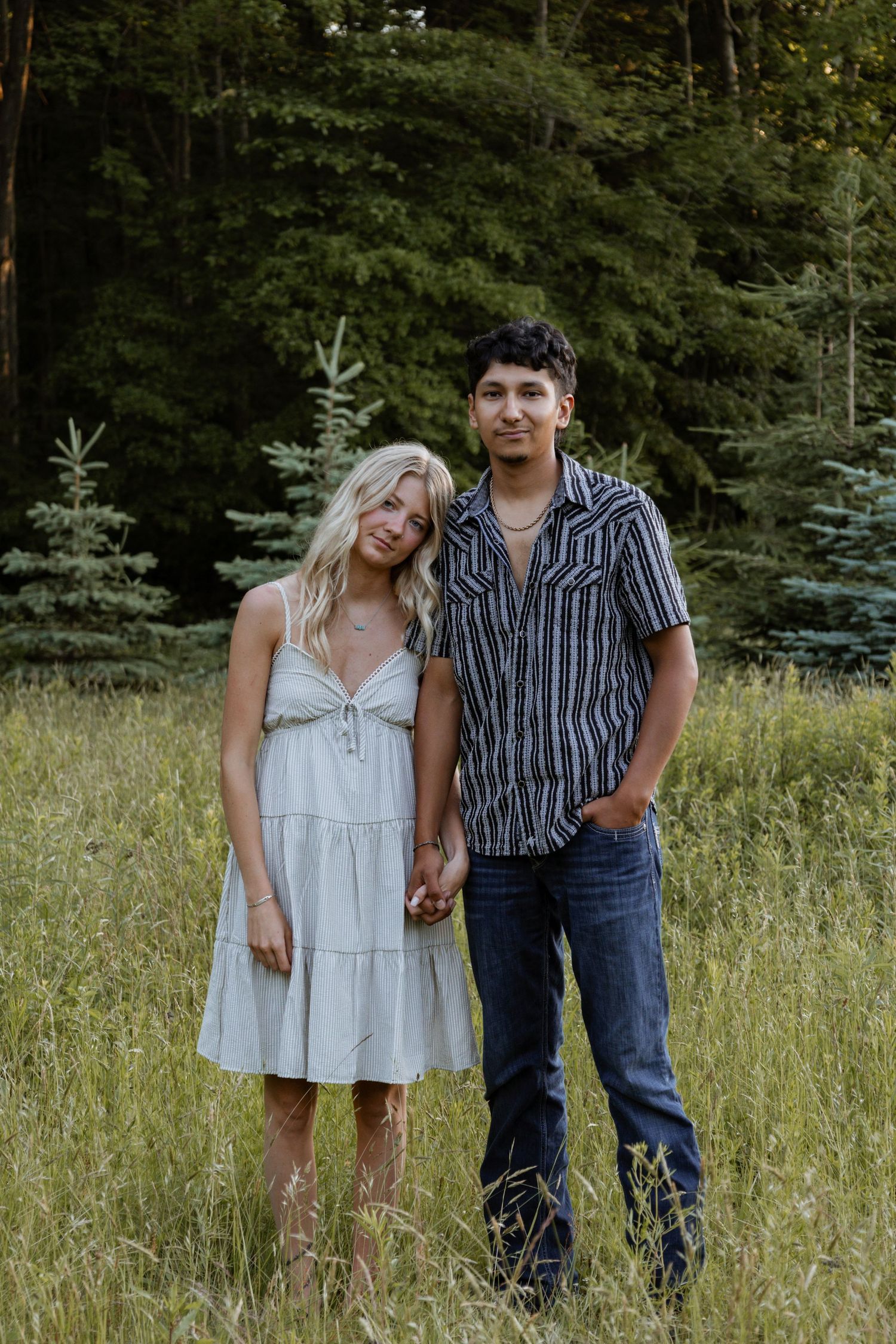 Couple holding hands in tall grass field with evergreen forest background.