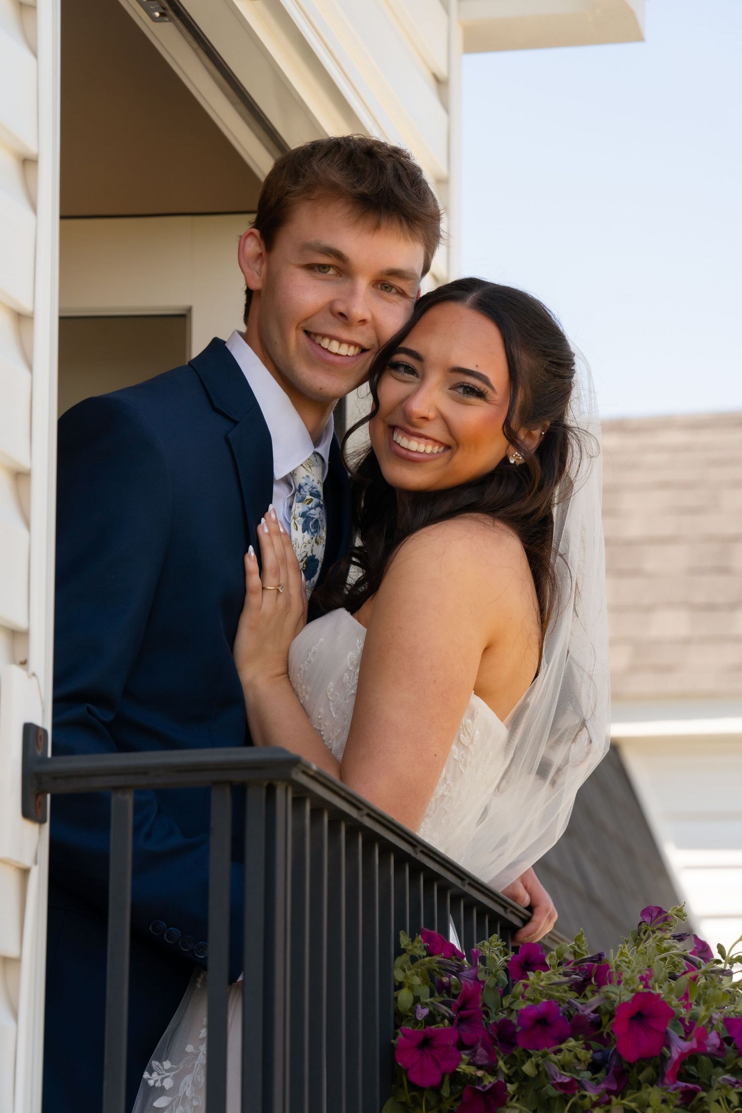 Smiling bride and groom pose together on a balcony adorned with purple flowers on their wedding day.