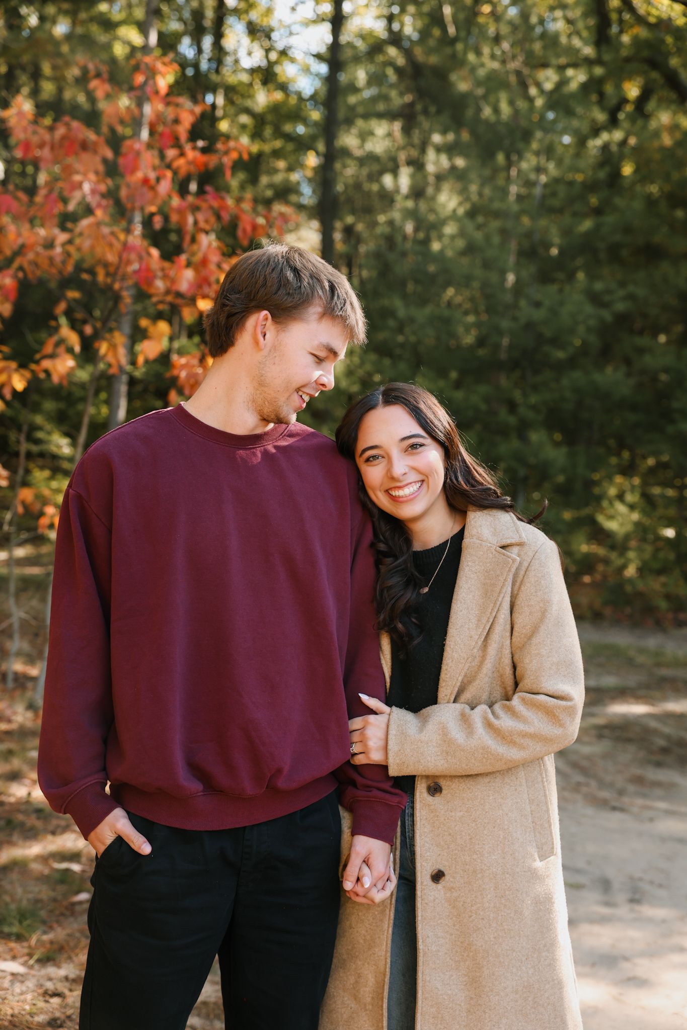 Smiling couple holding hands on autumn forest path, woman in beige coat.