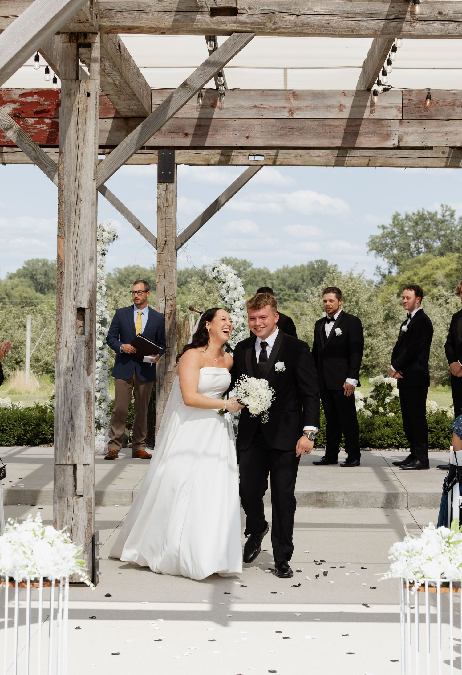 Bride and groom walk down the aisle at an outdoor wedding ceremony under a rustic wooden pergola.