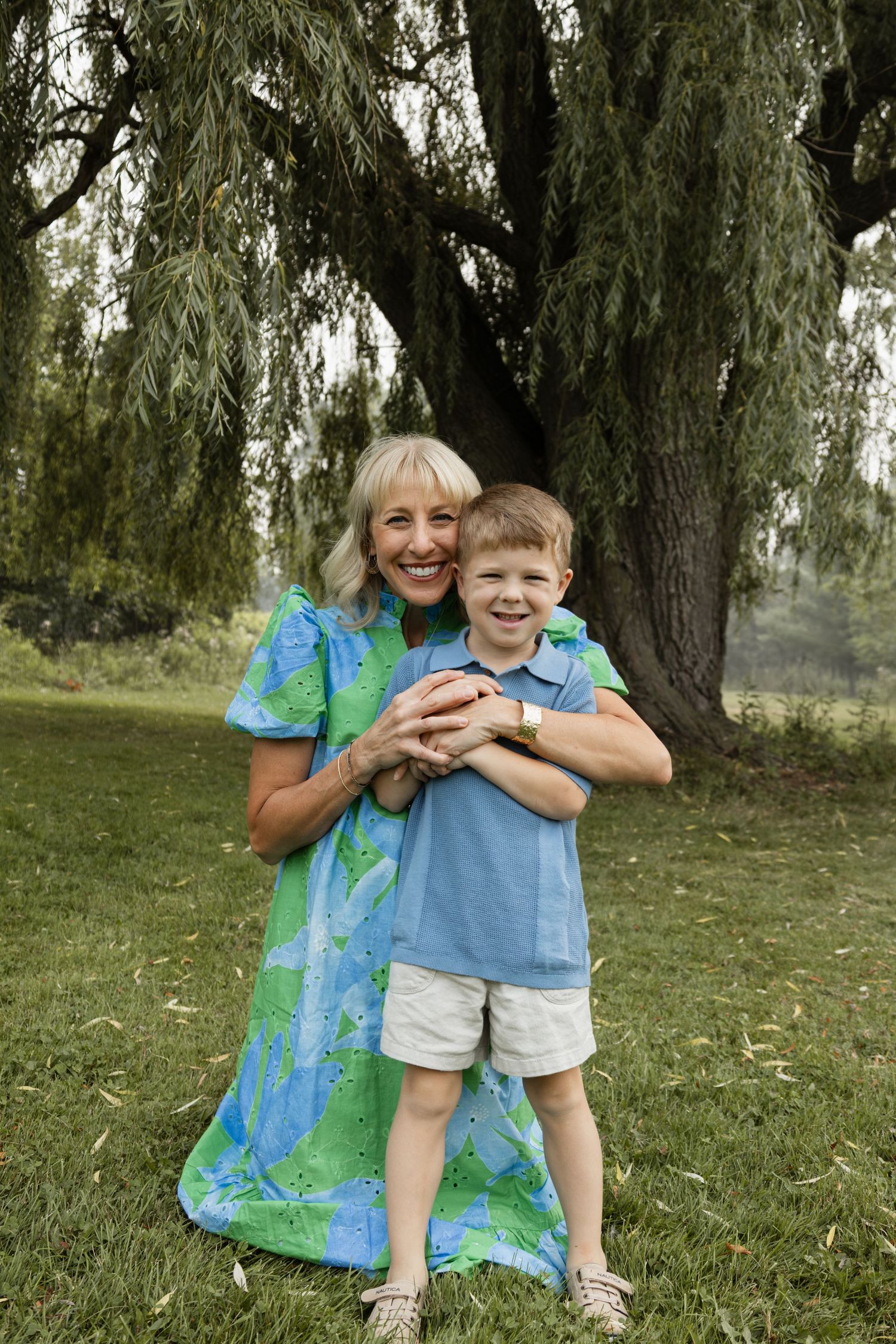 Smiling grandmother hugging young boy outdoors near willow tree on green grass.