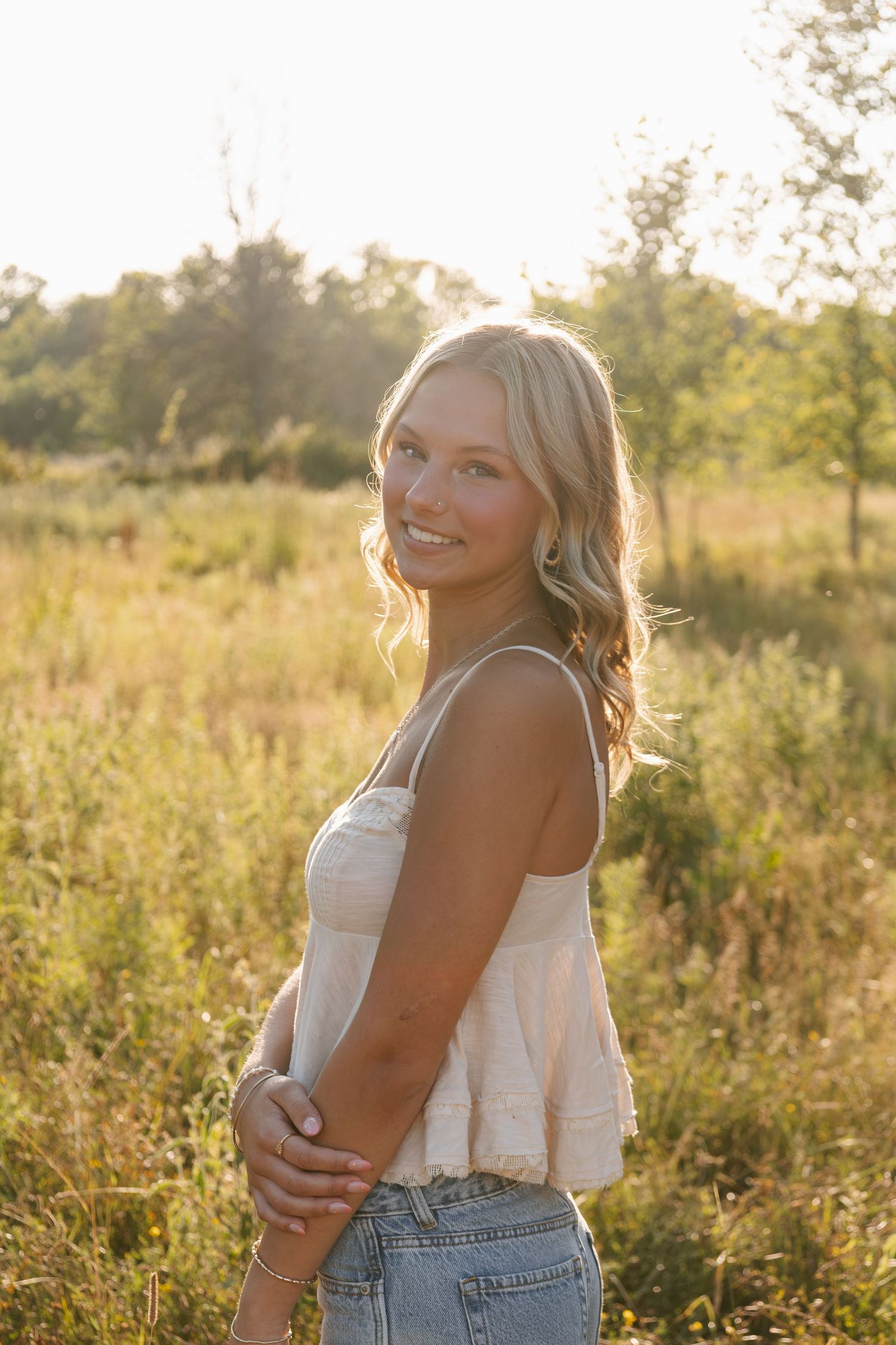 Young blonde woman smiling in golden hour sunlight, wearing white crop top and jeans in a lush green meadow.