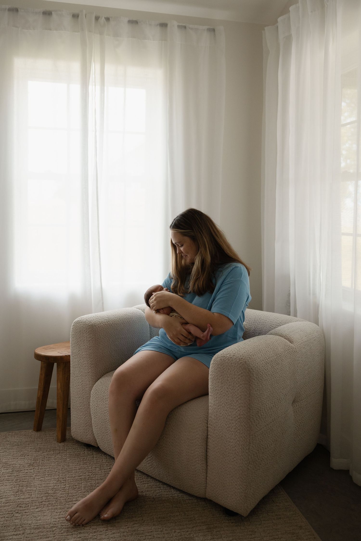 A mother in a blue dress sits in a beige armchair by sheer curtains, tenderly cradling her newborn baby.