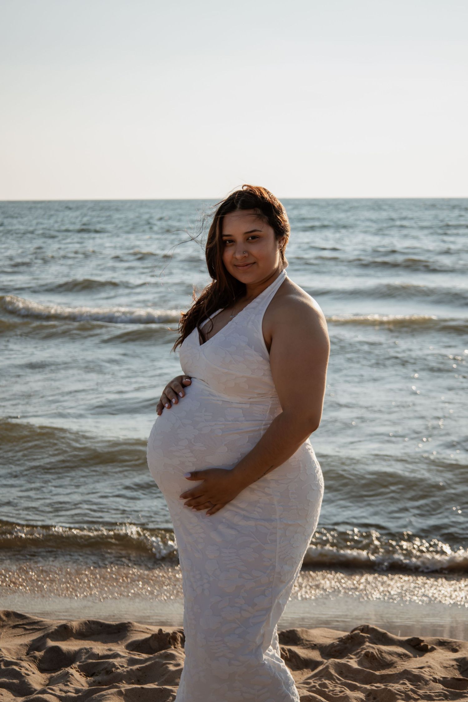 Pregnant woman in white lace dress cradling baby bump on sandy beach with ocean waves in background.