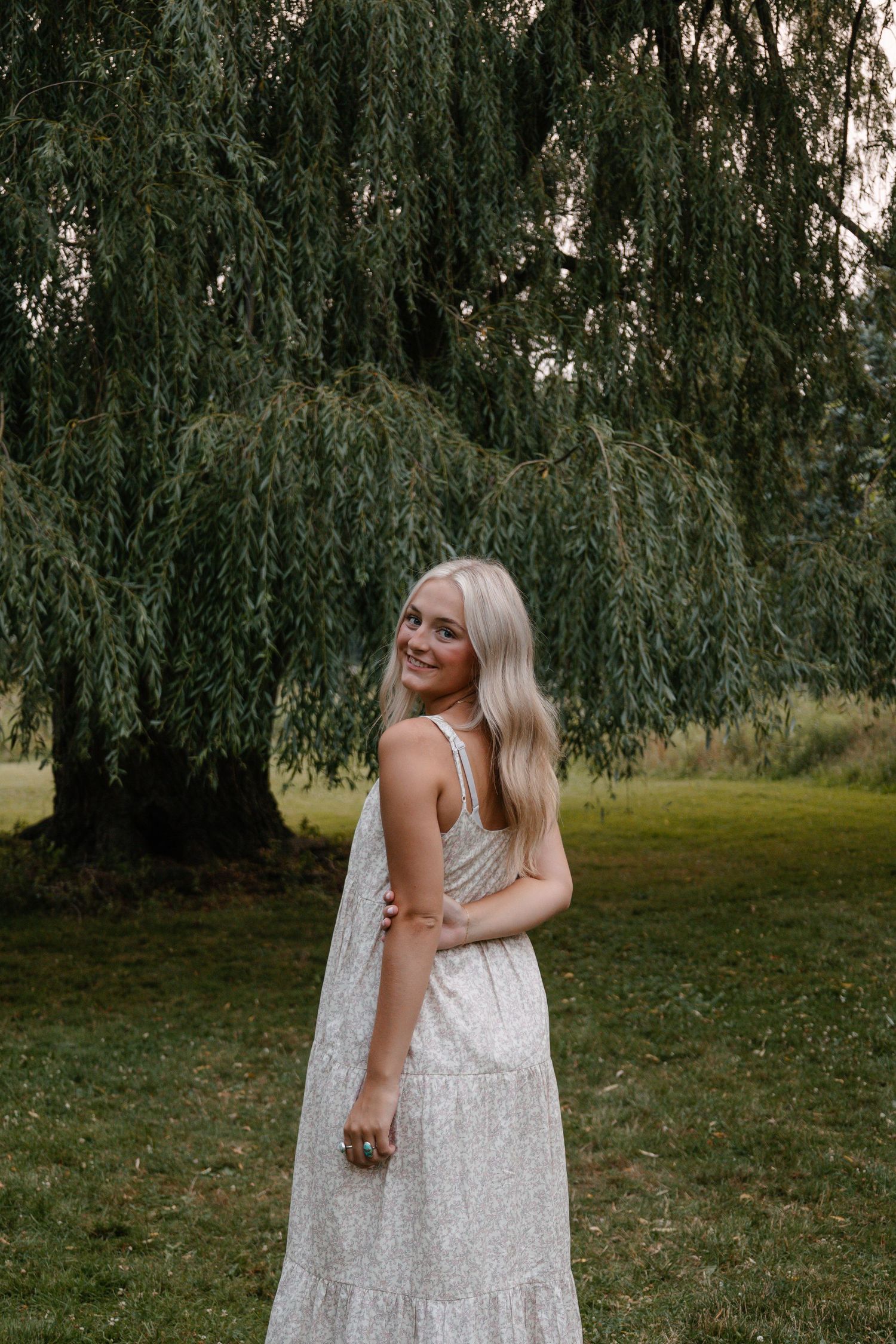 Smiling blonde woman in white floral maxi dress posing gracefully under a weeping willow tree.