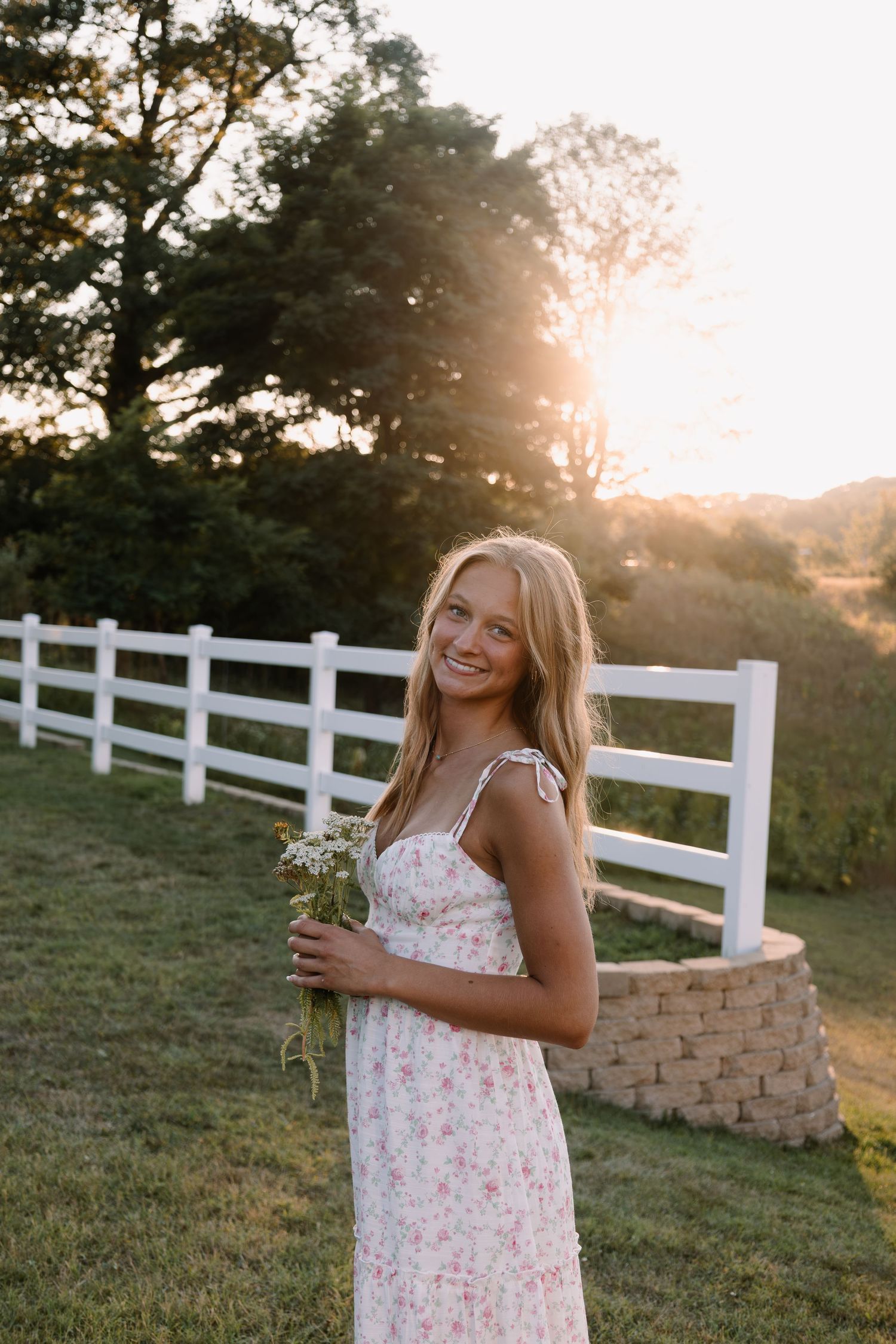Young woman in floral sundress holding wildflowers, smiling at sunset near white fence in countryside.