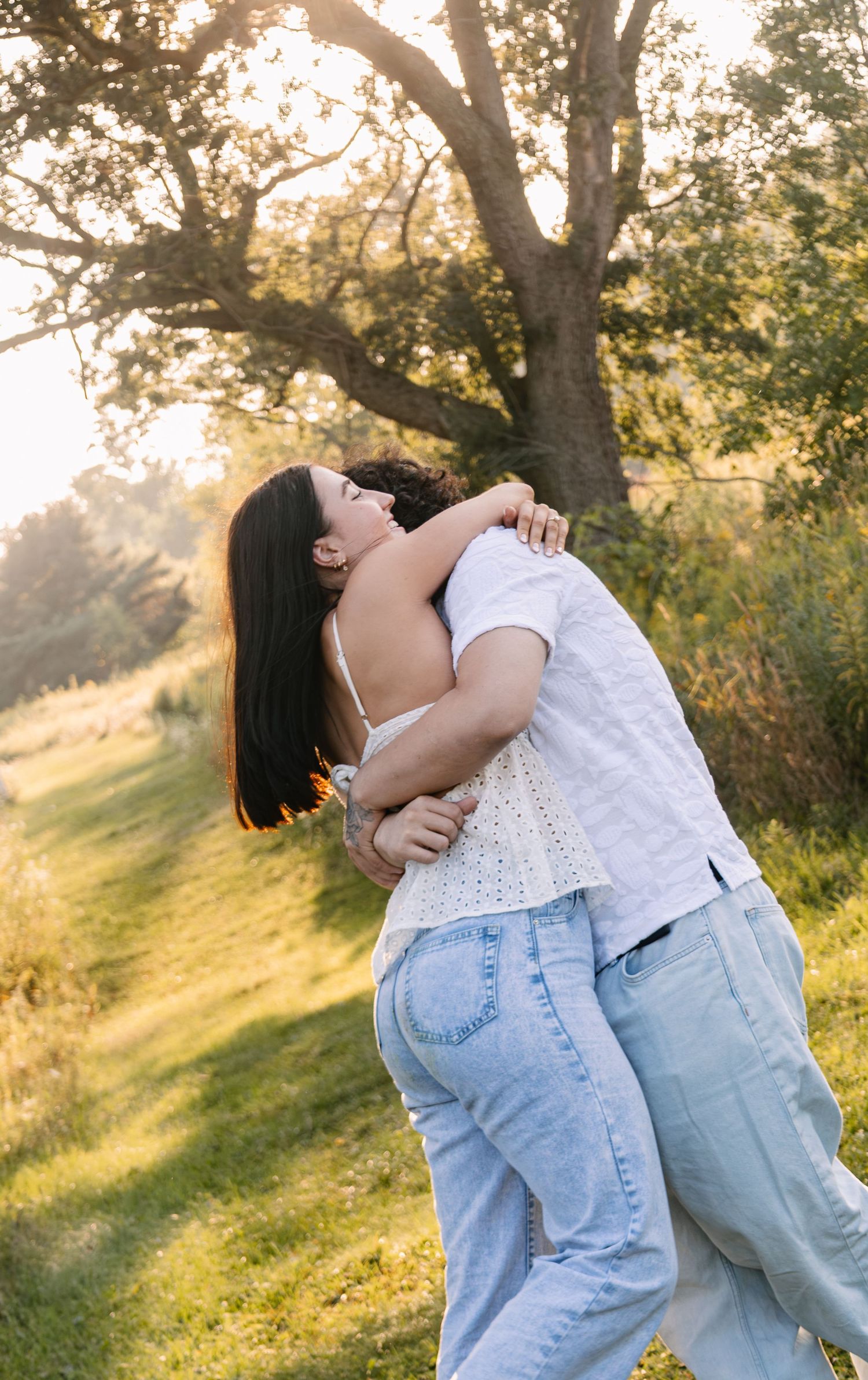 Couple embracing and kissing outdoors in golden hour sunlight near a large tree on a green hillside.