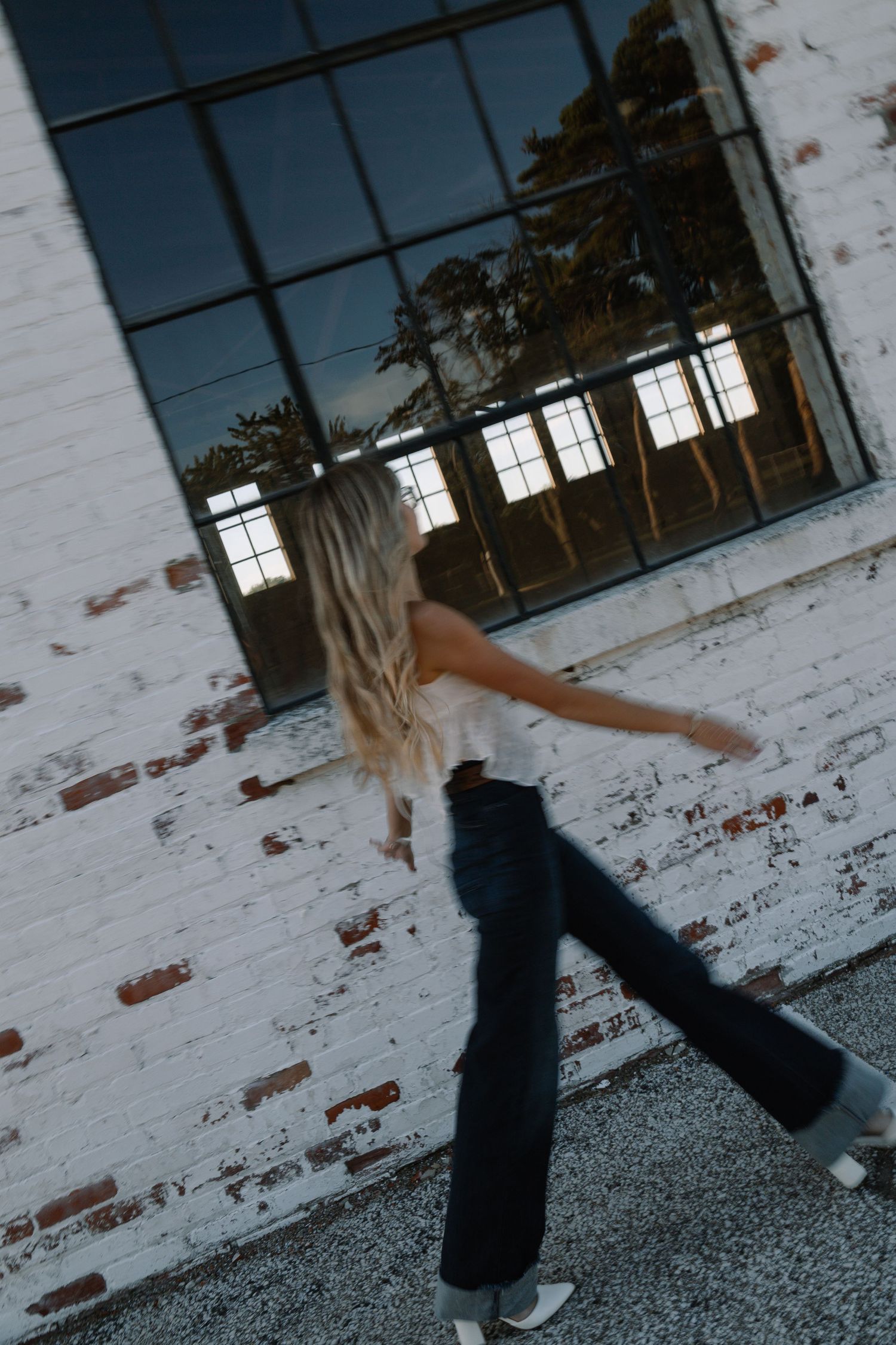 Blonde woman in wide-leg jeans and white top posing dynamically against a white brick wall with large black windows.