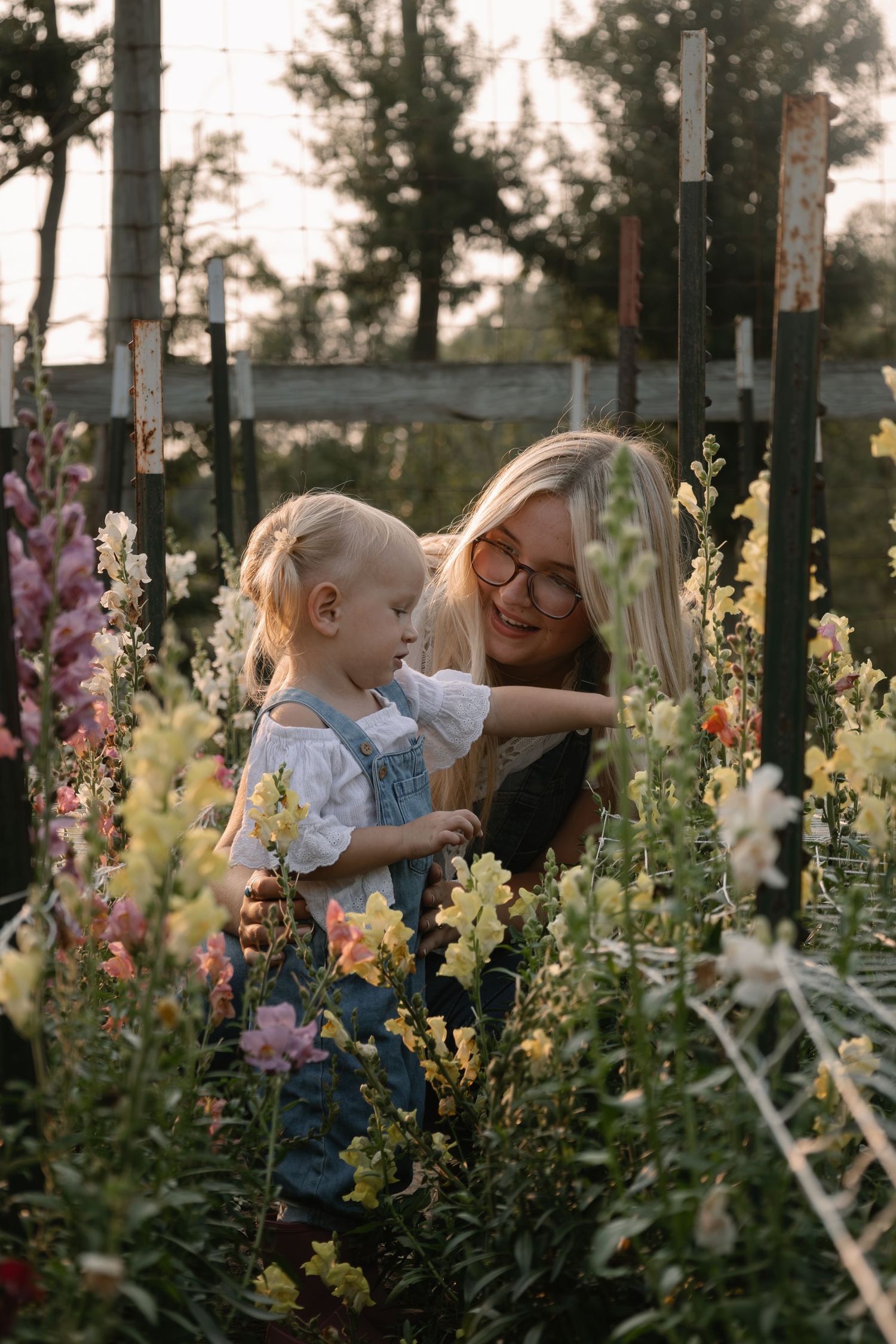 A smiling blonde woman with glasses holds a toddler girl among colorful snapdragons in a sunny garden.