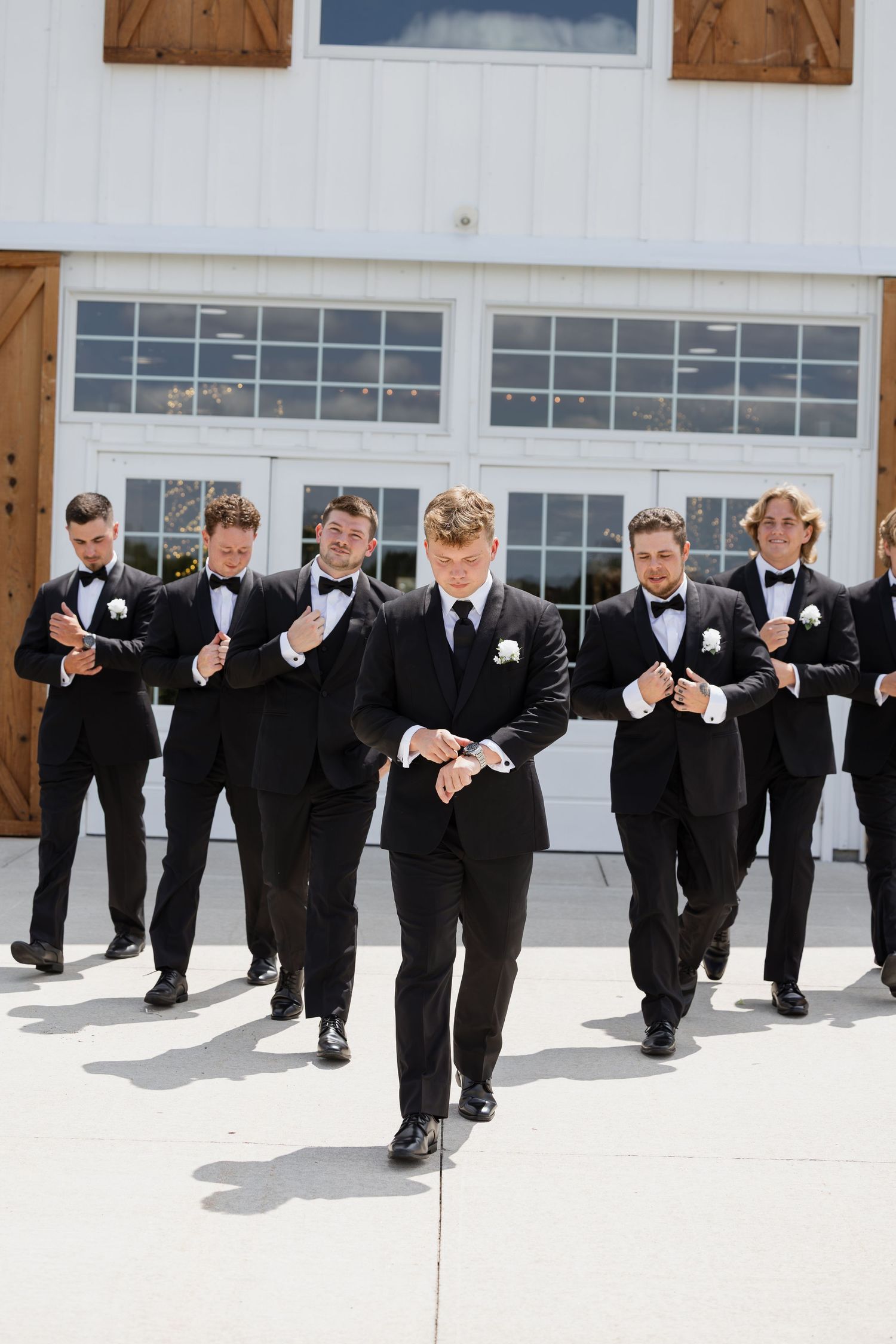 Groom leads groomsmen in black tuxedos walking confidently outside a white barn wedding venue.