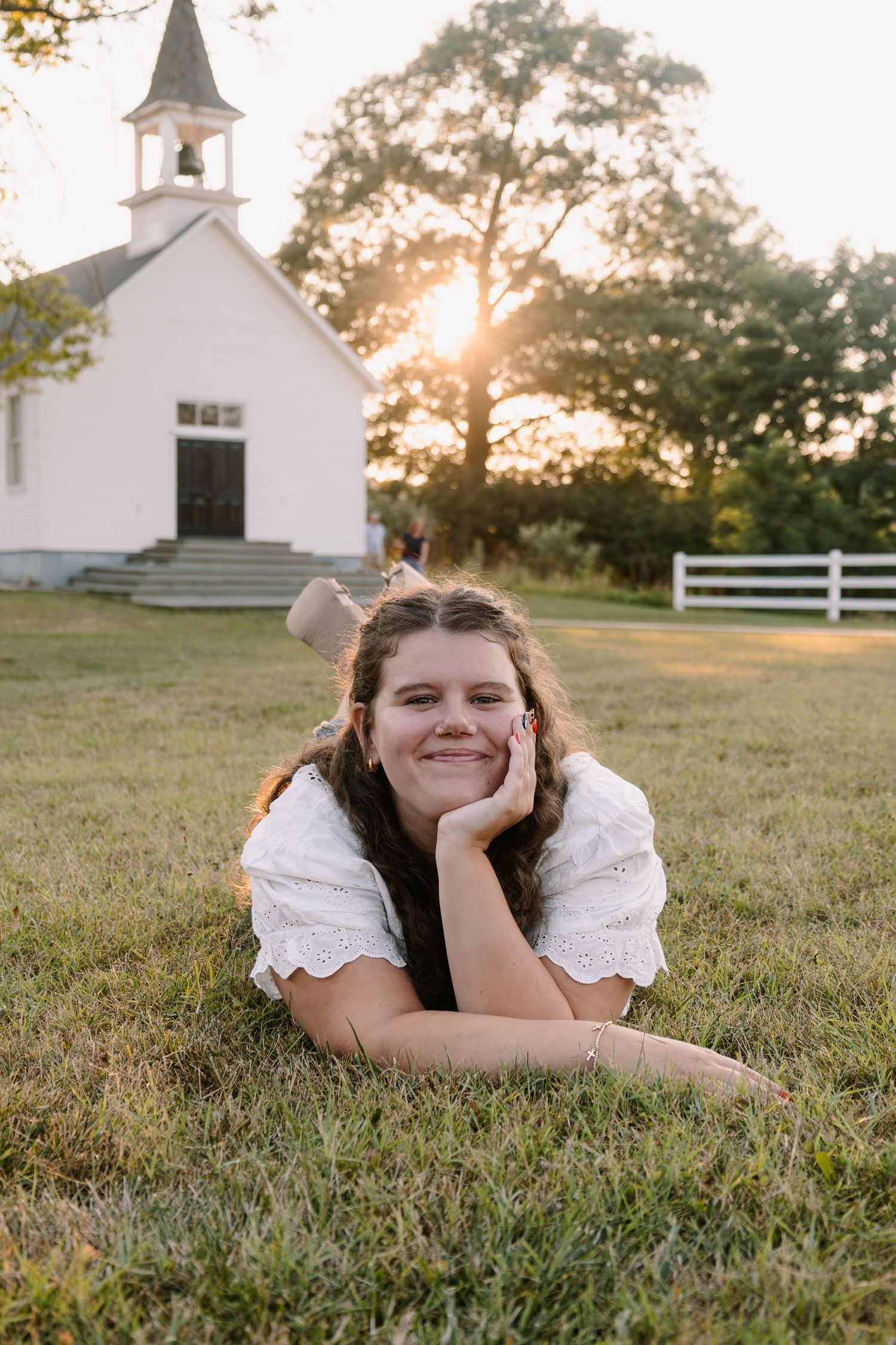 Young woman in white dress lying on grass smiling at sunset with a white country church in the background.