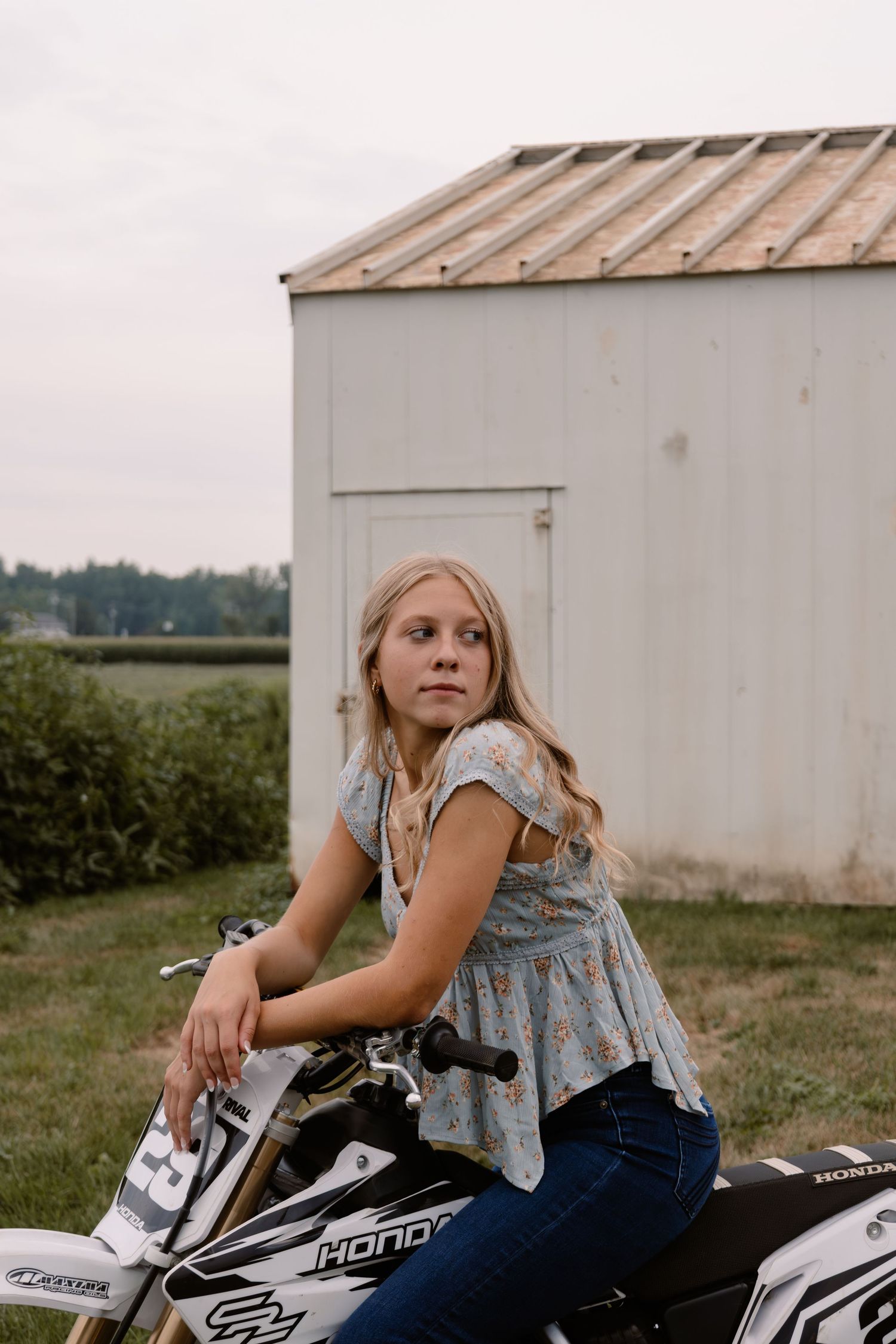 Young blonde girl sitting on a white Honda dirt bike in front of a shed, wearing a floral top and jeans.