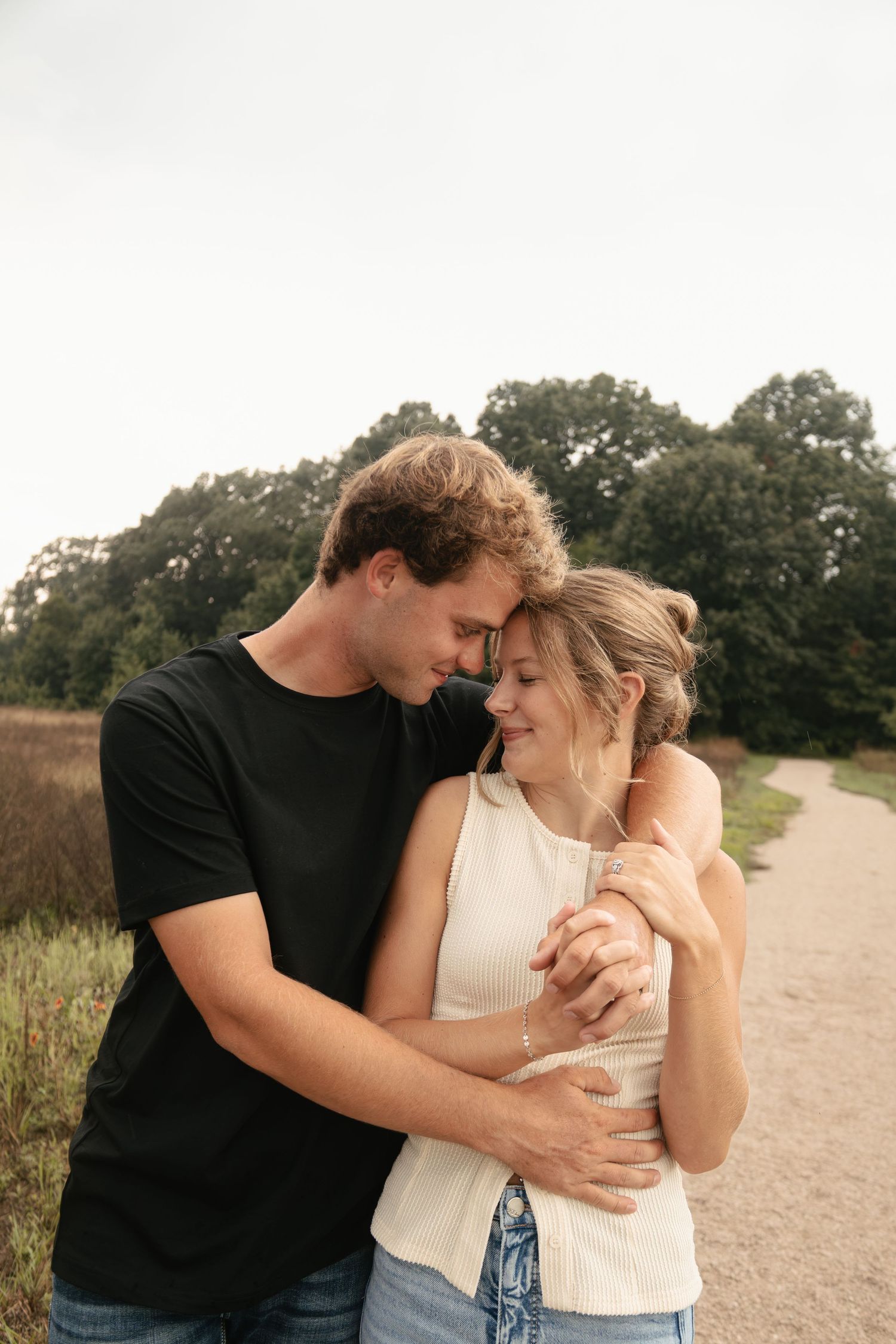 A young couple embraces tenderly on a rural path, surrounded by lush green trees and fields on a cloudy day.