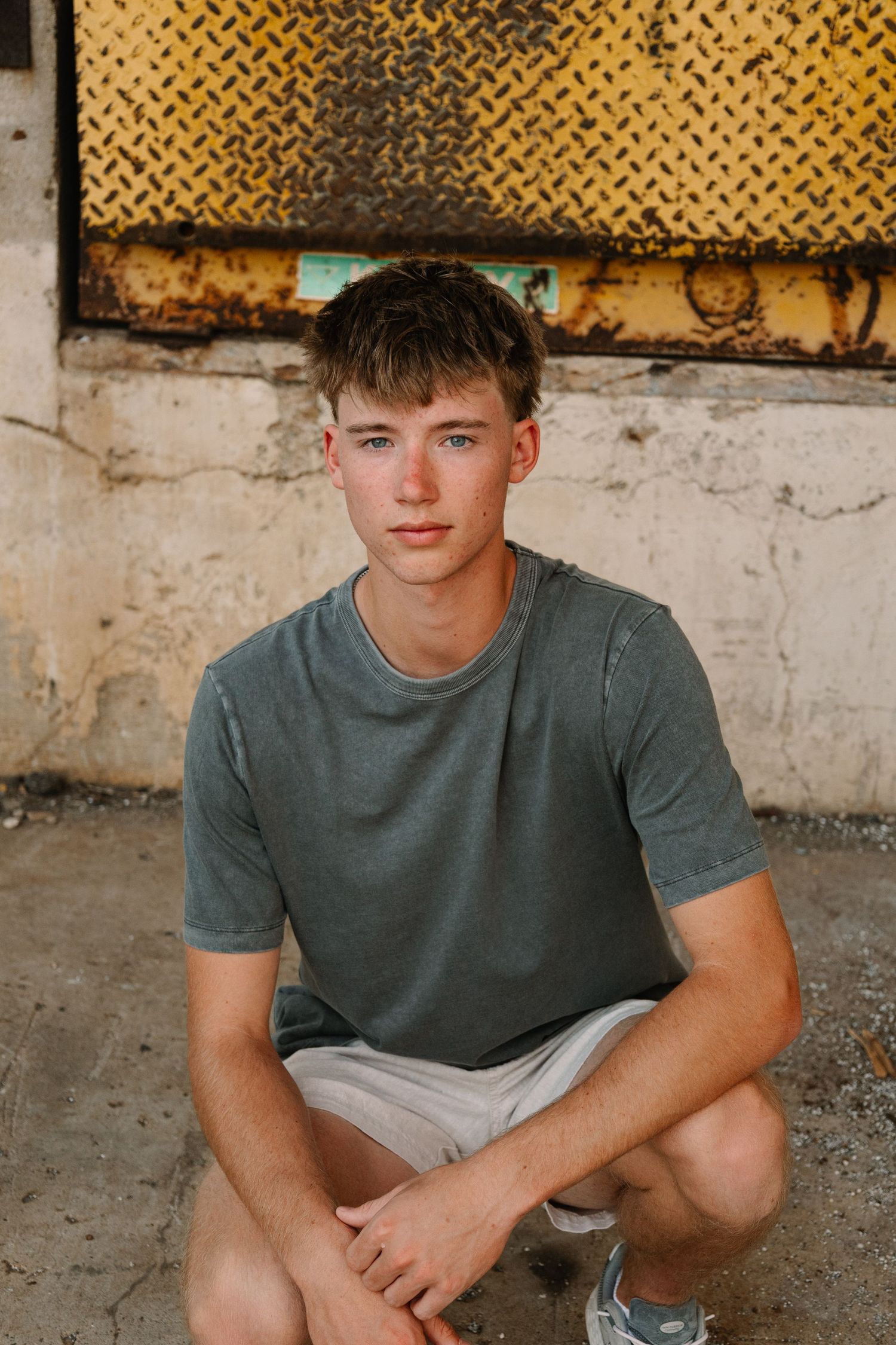 Young man in gray t-shirt and shorts crouching against rustic yellow industrial wall outdoors.