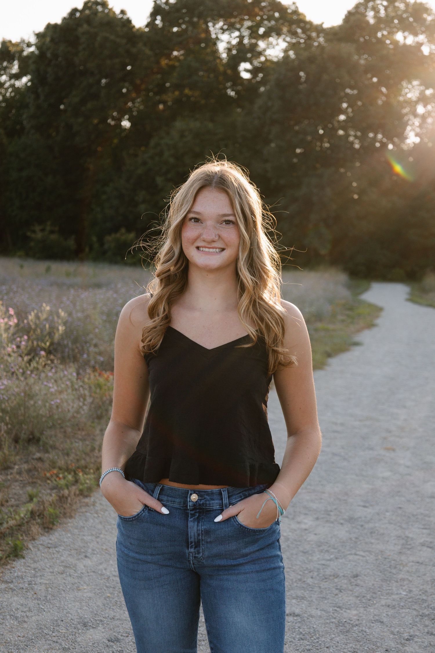 Young woman in black tank top and jeans smiling on a sunlit dirt path surrounded by trees.