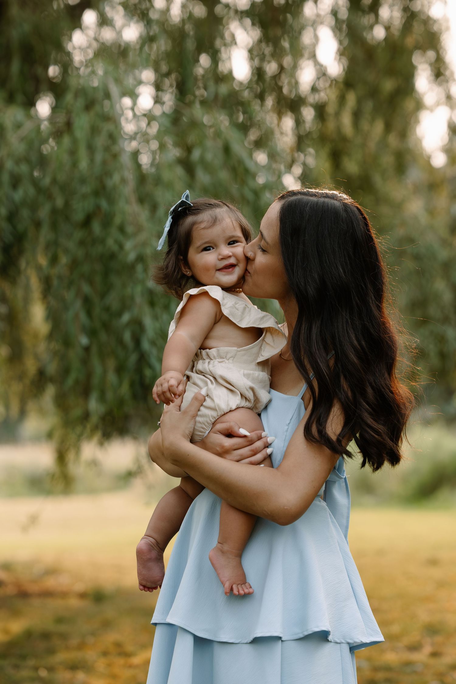 A mother in a blue dress kisses her smiling baby girl outdoors near a weeping willow tree.