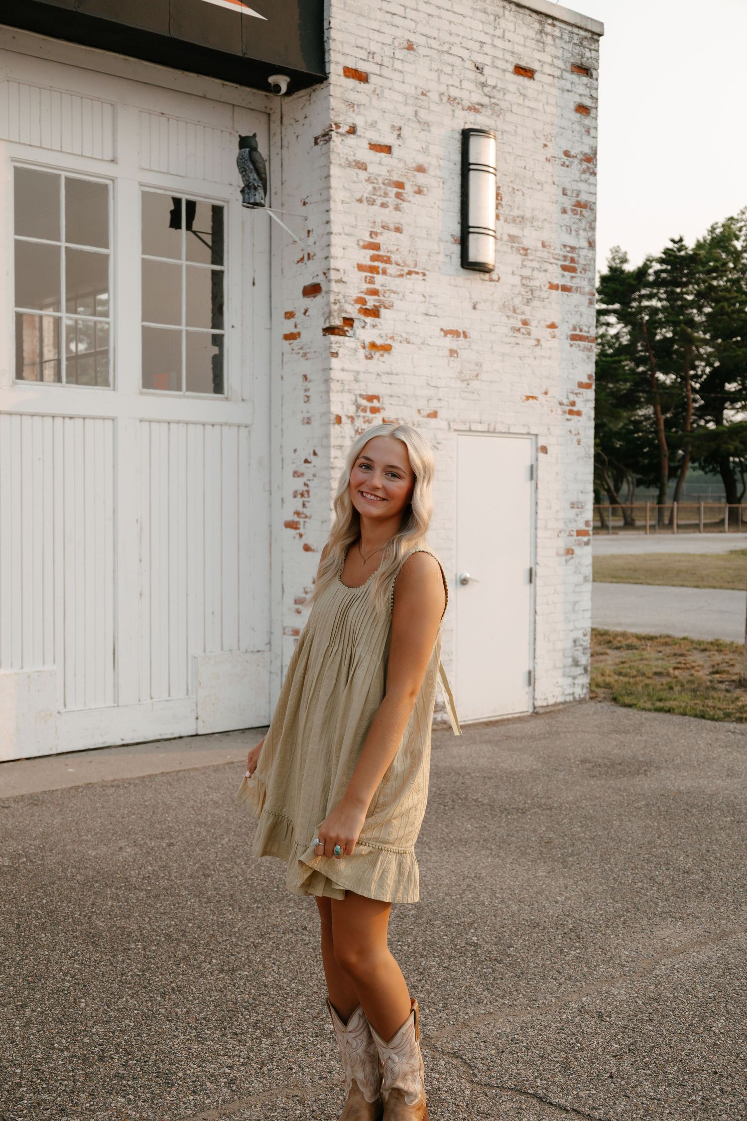 Young blonde woman in sage green mini dress and cowboy boots smiling outside a white brick building.