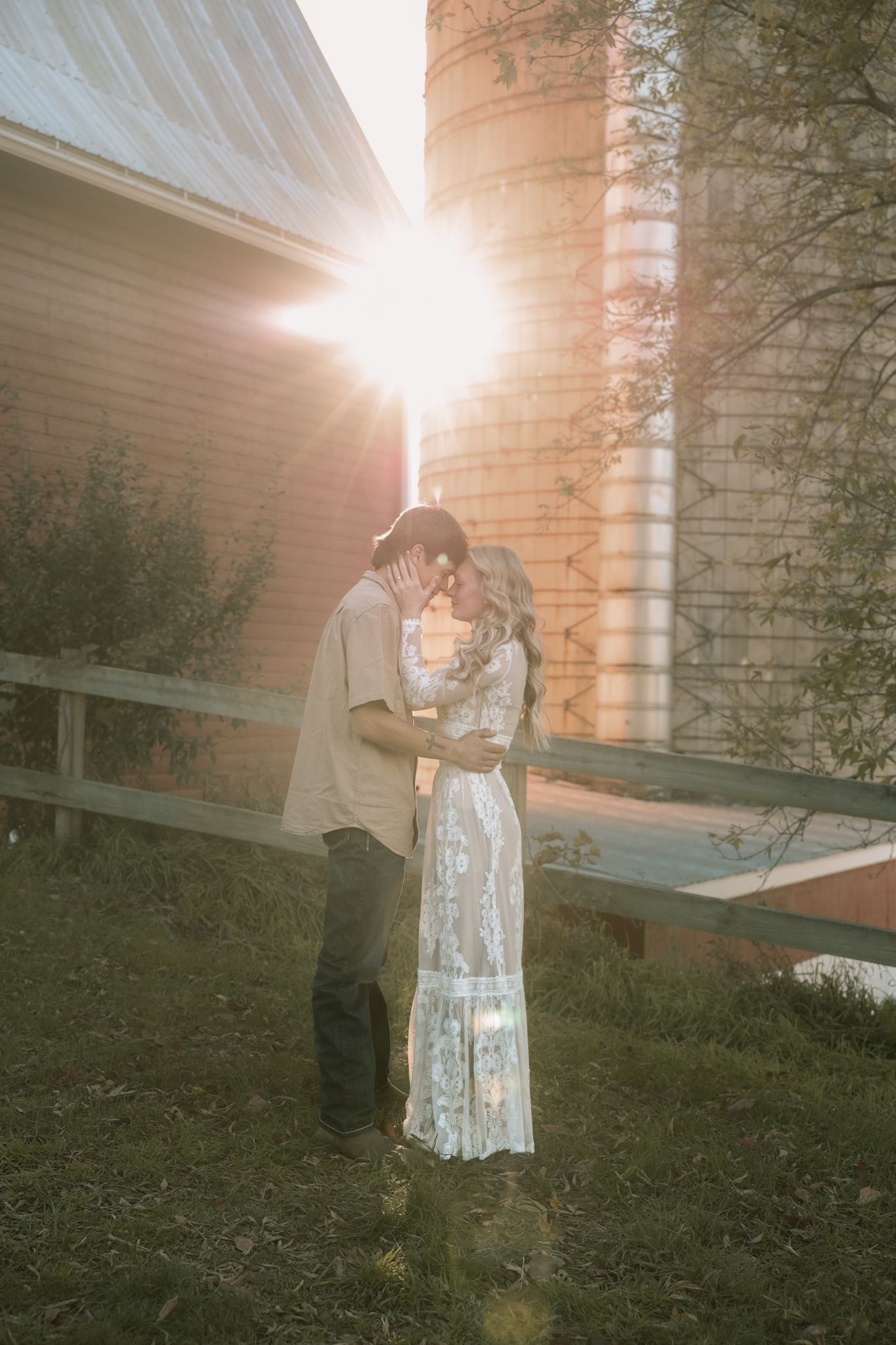 Couple kissing at sunset with golden lens flare between rustic grain silos on a farm.