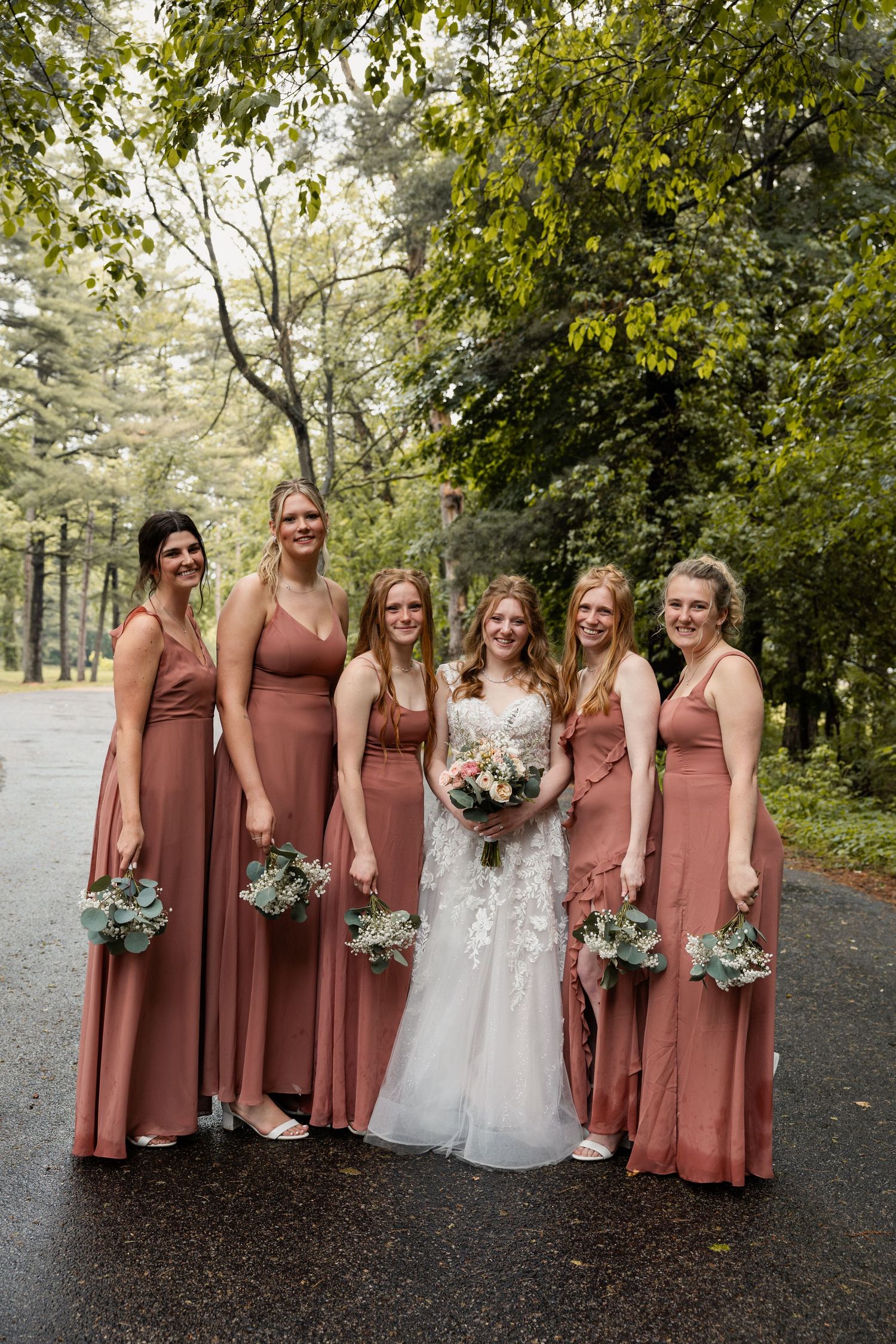 Bride poses with five bridesmaids in mauve dresses holding bouquets on a tree-lined path.