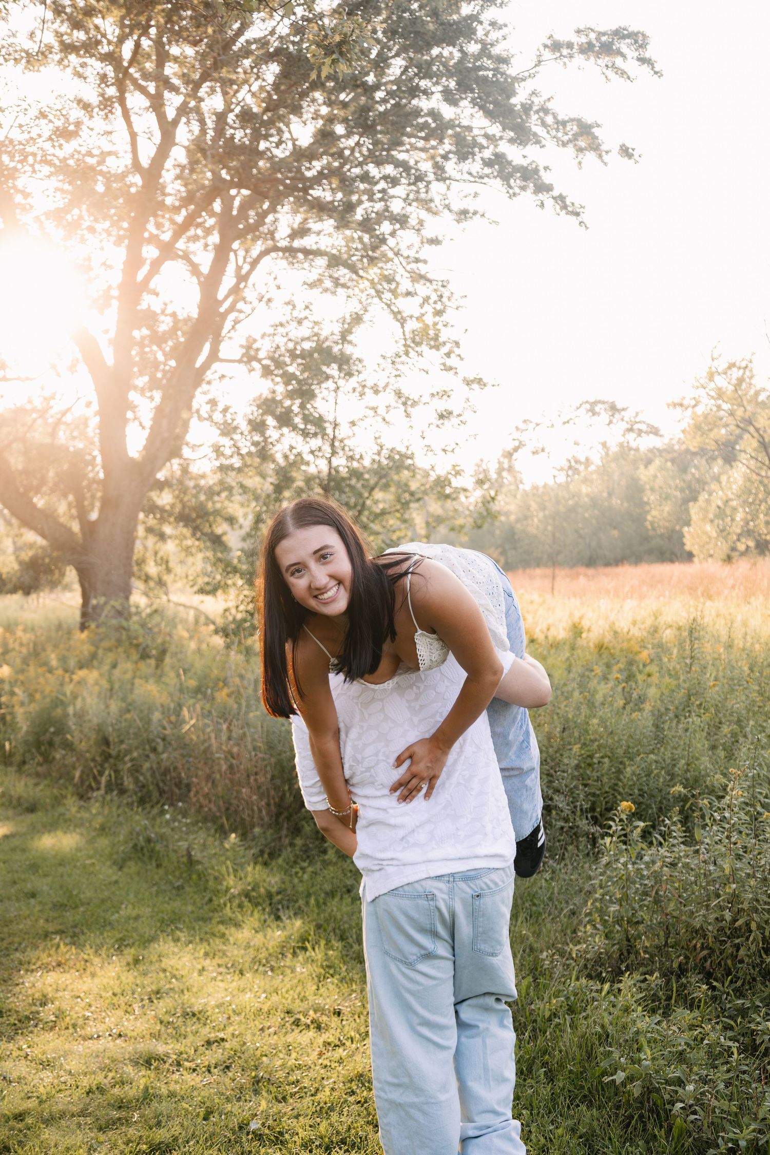 Young woman smiling outdoors in golden hour sunlight, wearing white tank top and light blue jeans in a green field.