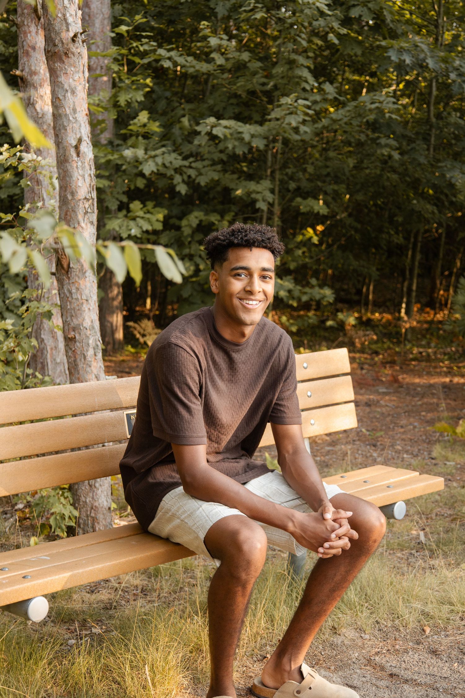 Young Black man smiling on a wooden park bench, wearing brown t-shirt and shorts, with green trees in background.