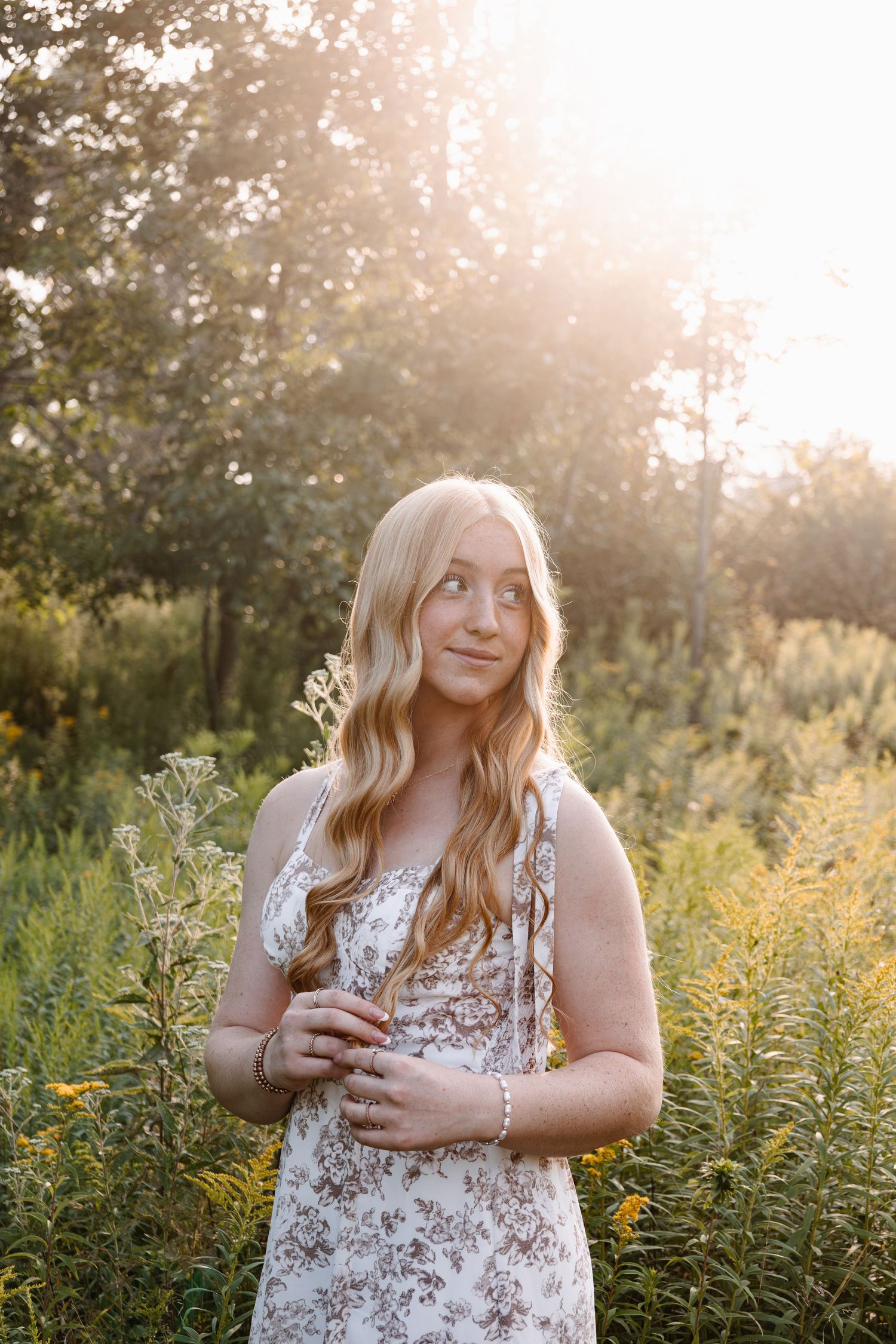 Young blonde woman in floral dress standing in golden sunlit meadow with wildflowers and trees in background.