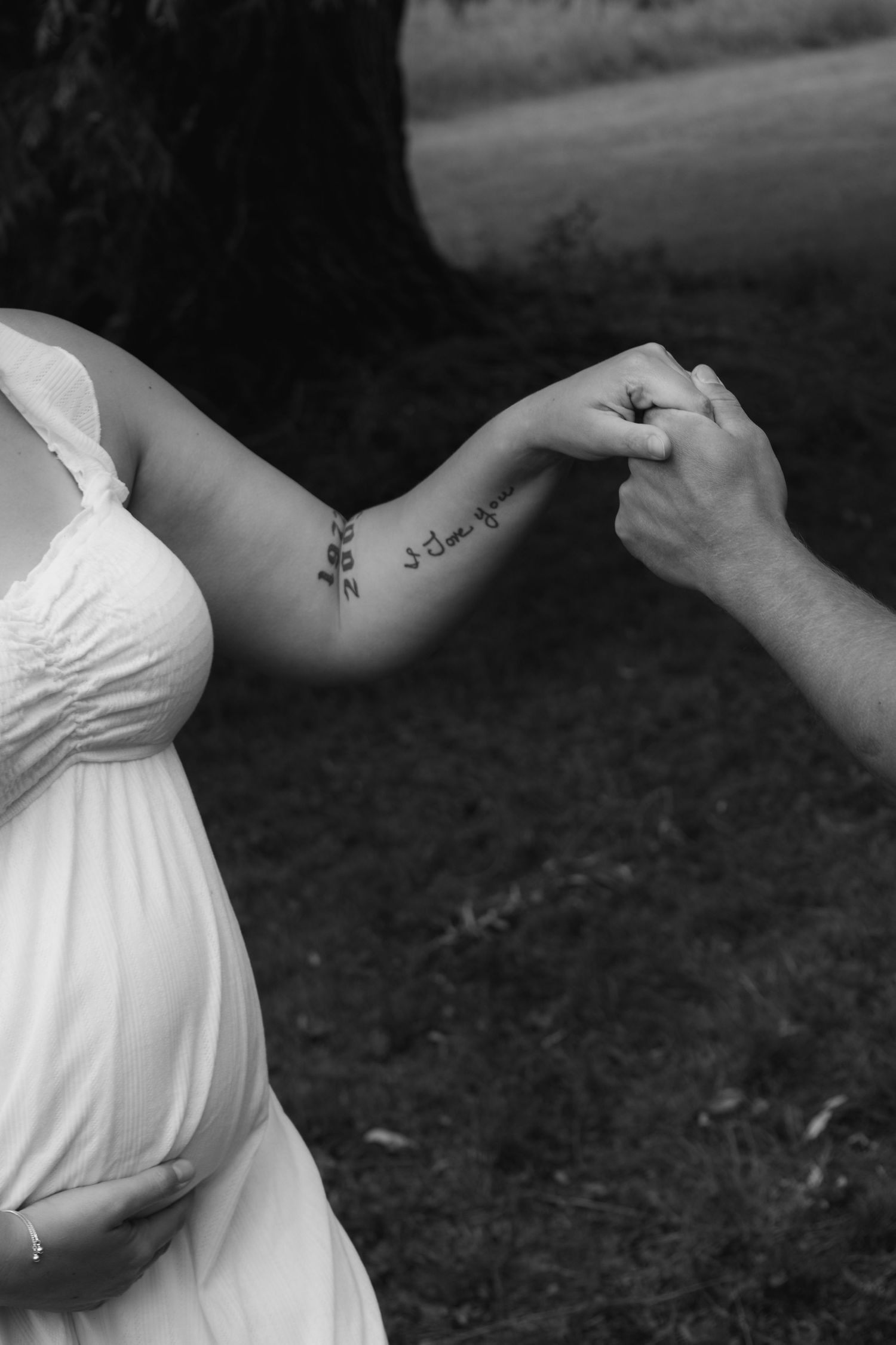 Black and white close-up of pregnant woman in white dress holding hands with partner, showing arm tattoo.