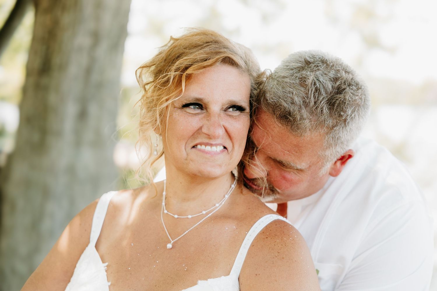 Happy bride smiles as groom whispers in her ear during romantic outdoor wedding portrait.
