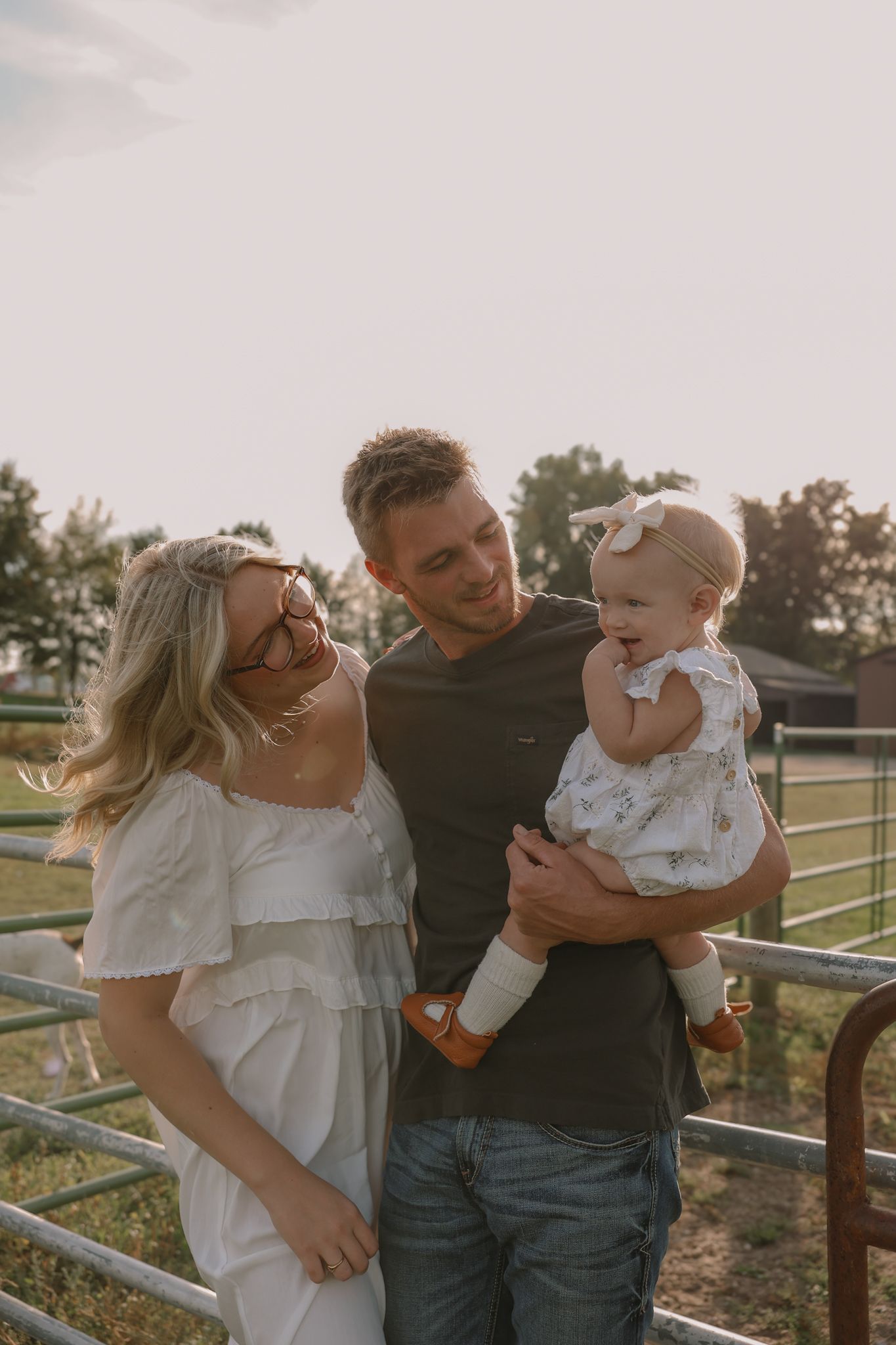 Young family smiles together at golden hour near a farm fence holding their baby girl.