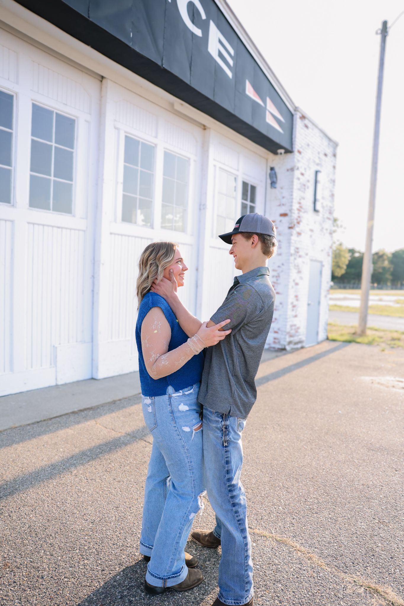 A couple stands facing each other outside a white building, the woman in a blue top and ripped jeans, the man in a gray shirt and cap.