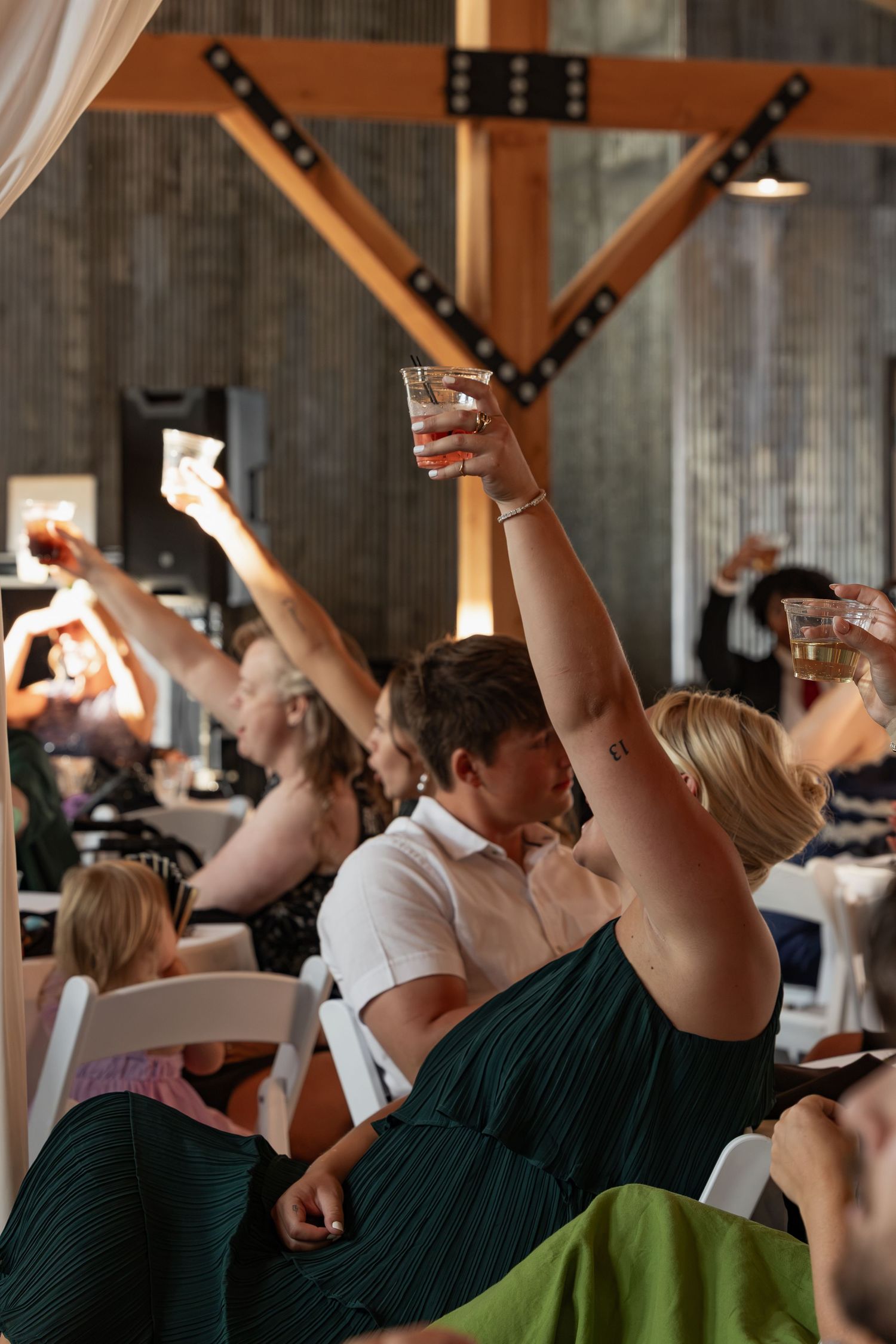 Wedding guests raising glasses in a toast inside a rustic barn reception venue with wooden beams.