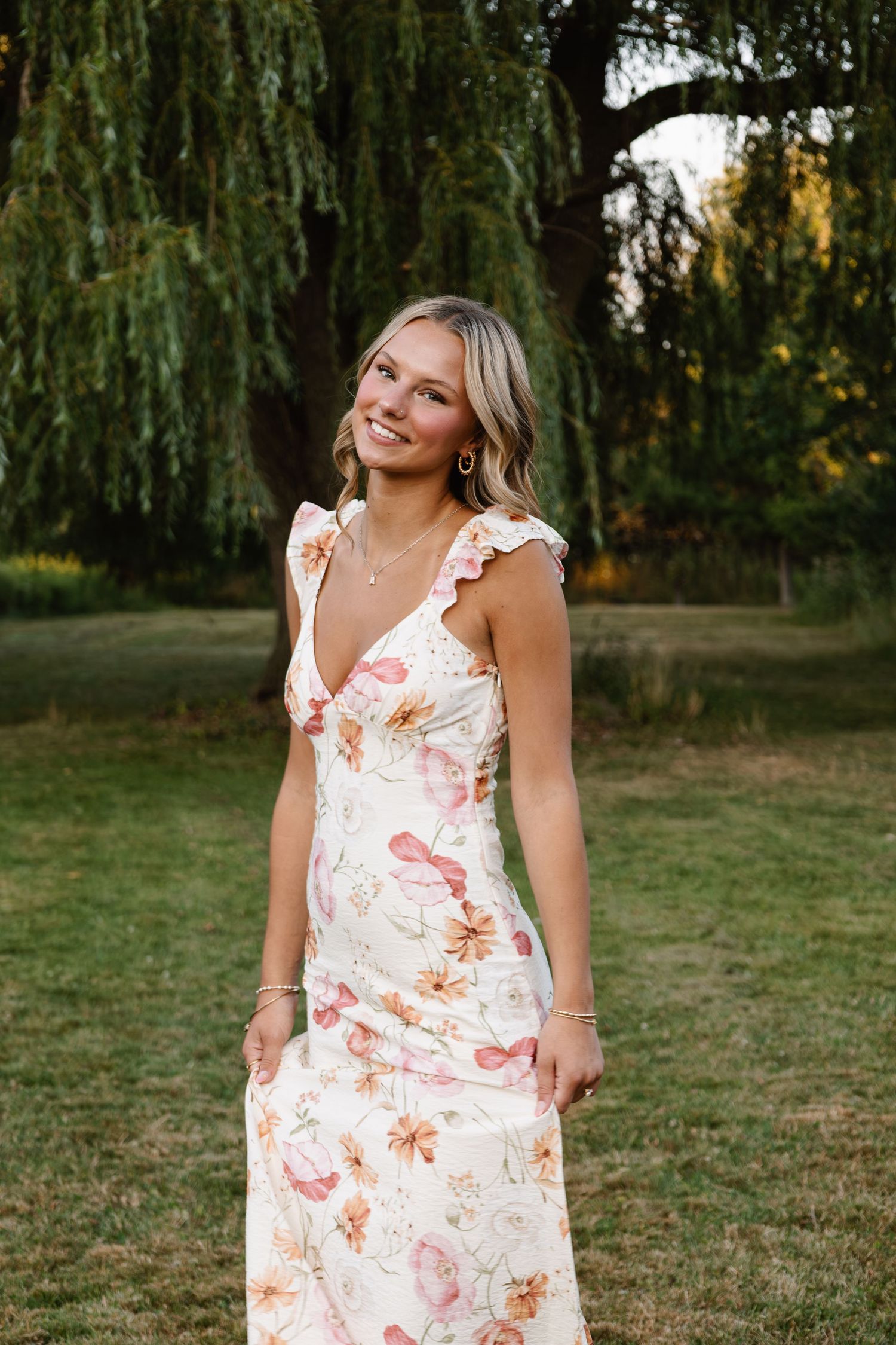 Young woman smiling in a floral maxi dress with ruffle straps, standing outdoors near a willow tree.