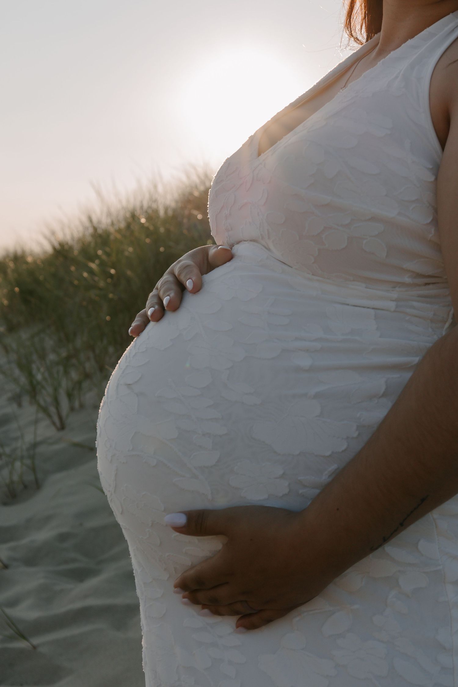Pregnant woman in white textured dress cradling her baby bump on a sandy beach at sunset.