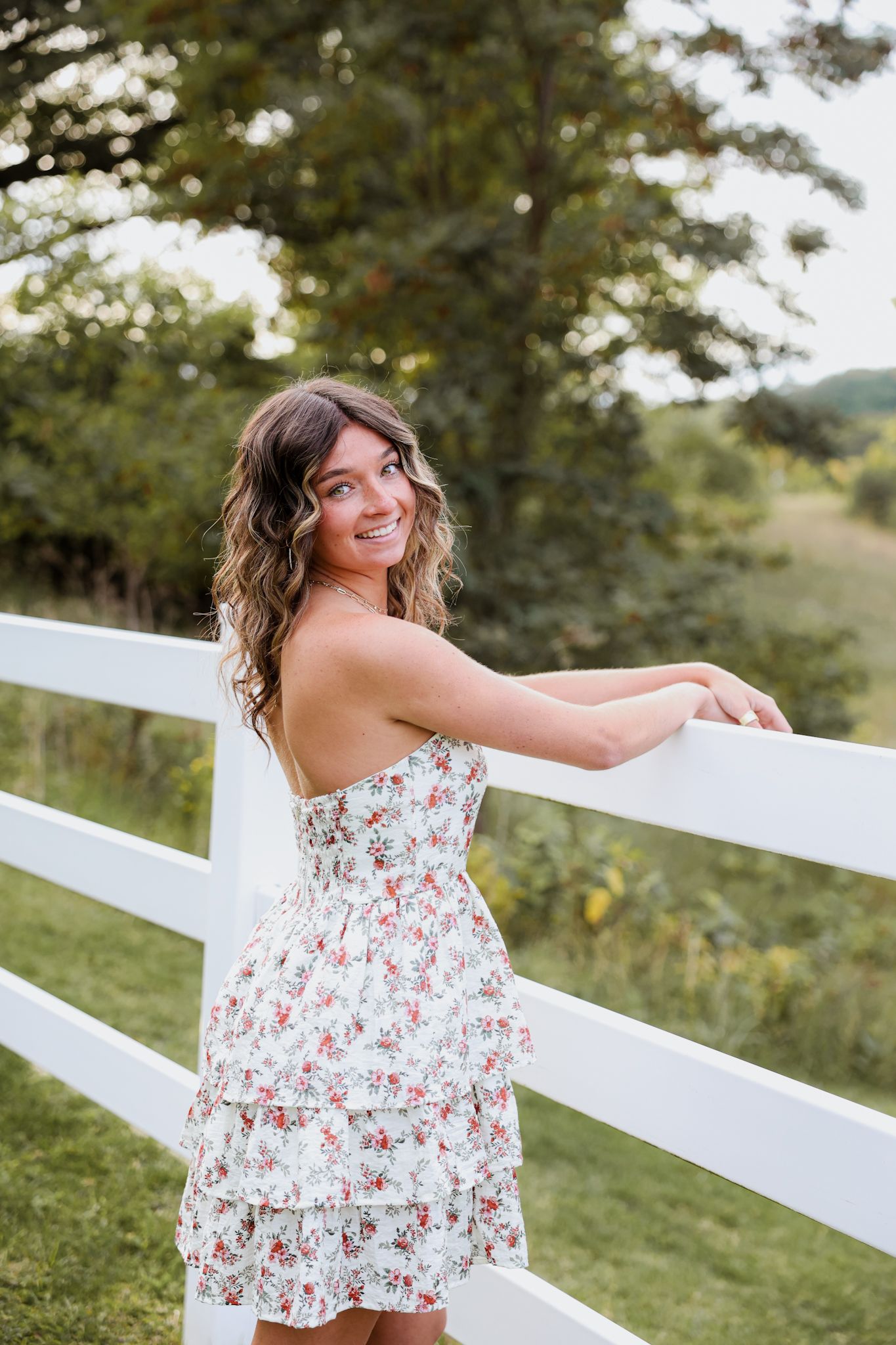A young woman in a floral tiered dress smiles by a white wooden fence outdoors.
