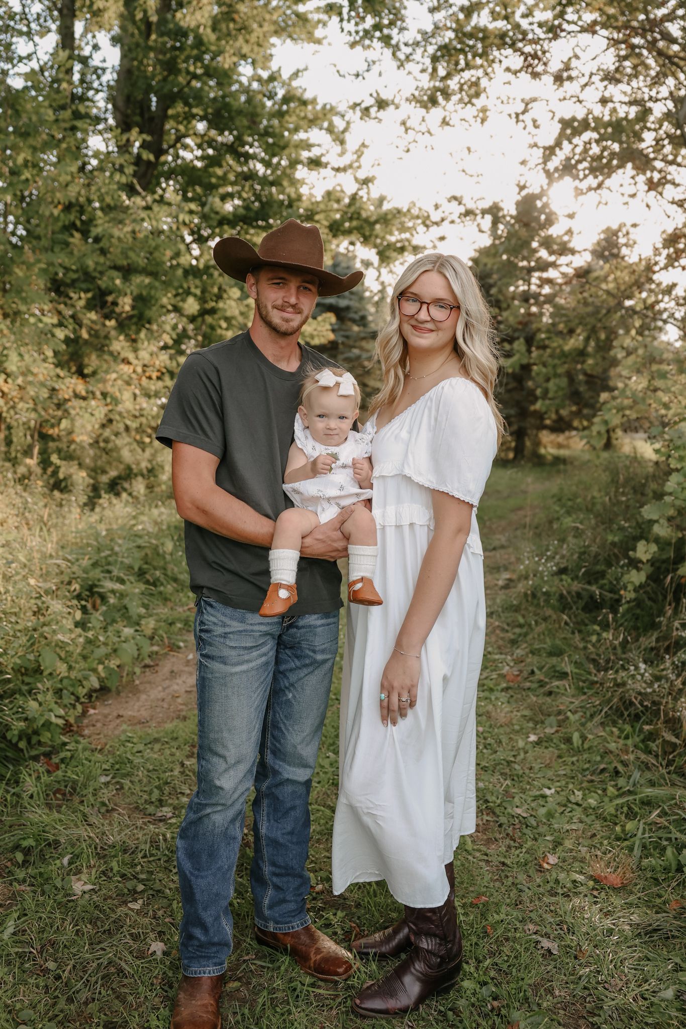 Young family posing on wooded path, dad in cowboy hat holding baby girl, mom in white dress.