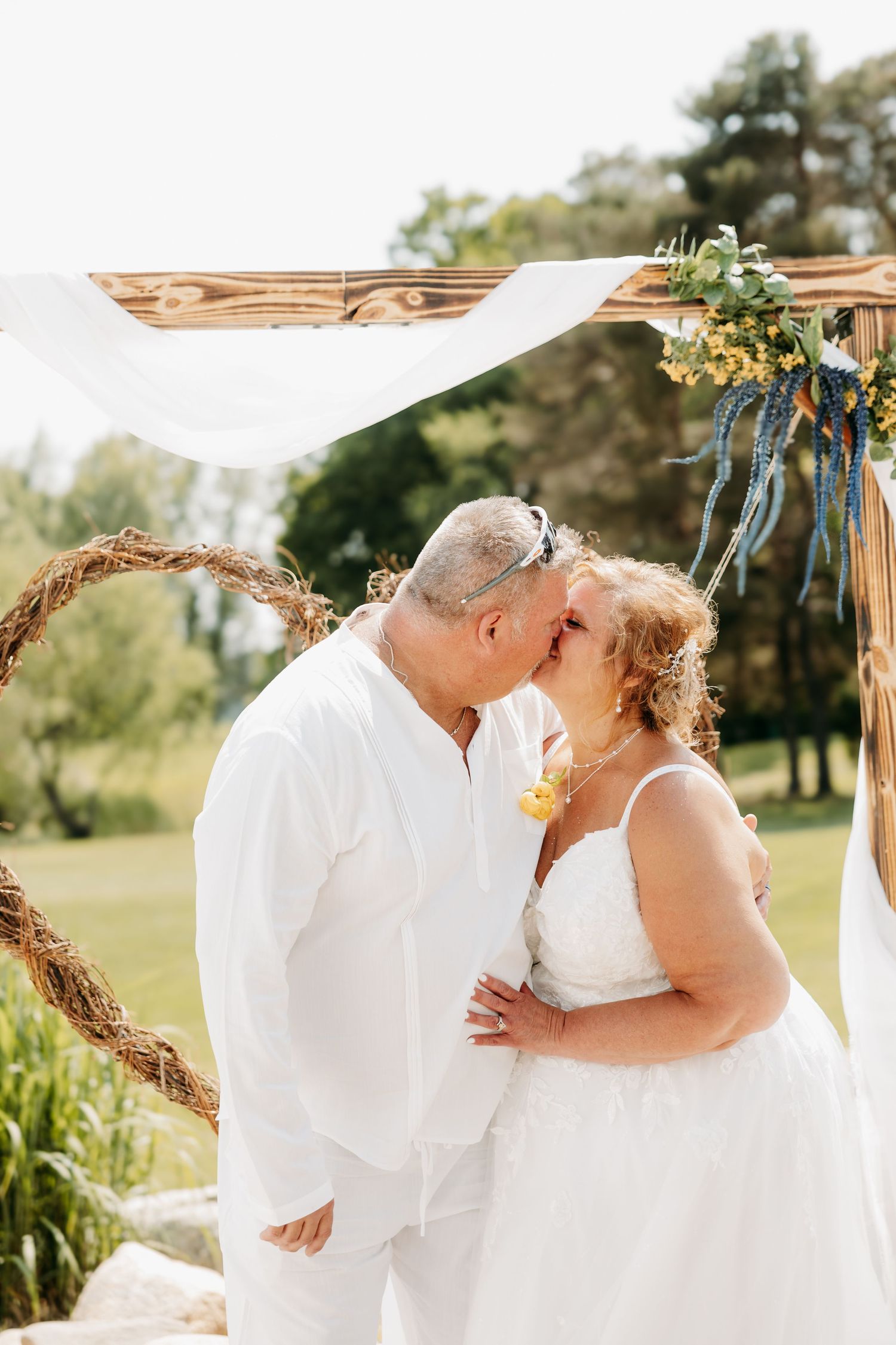 Mature couple shares first kiss as newlyweds under wooden arch with floral decorations outdoors.